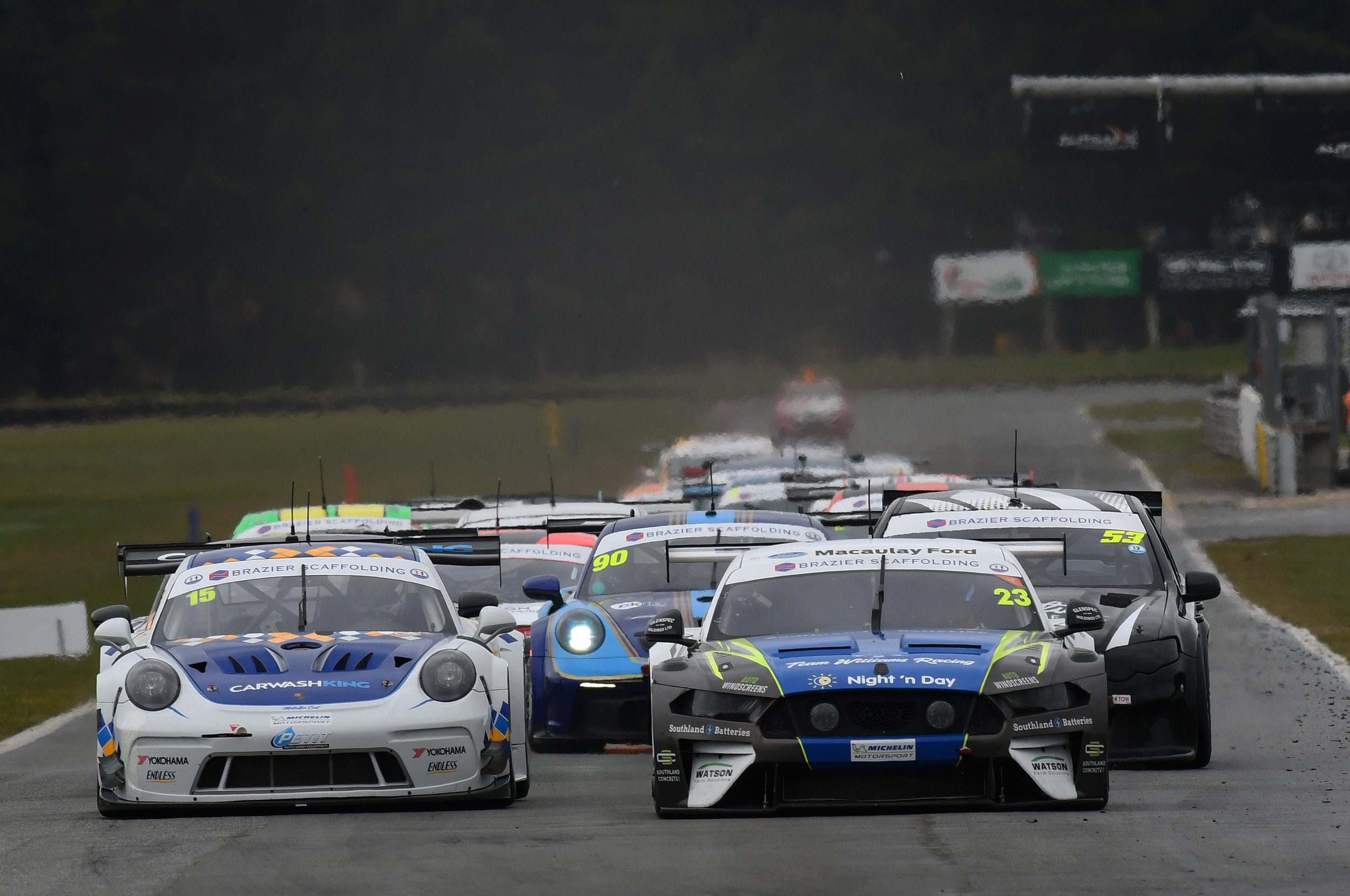 Multiple race cars on a race track during a race, with rain and wet conditions, some with sponsorship logos and numbers.