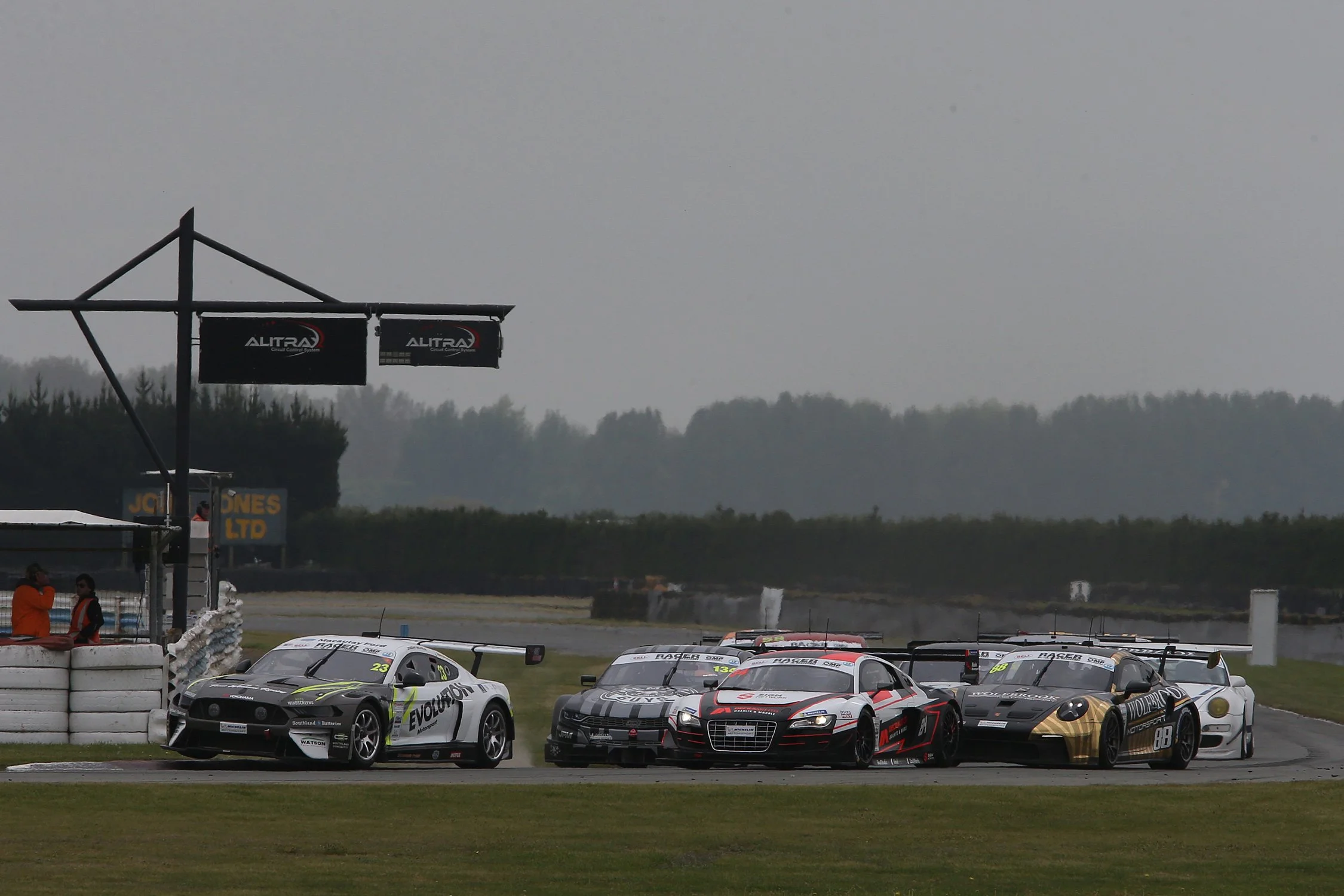 Race cars lined up on a racetrack under a cloudy sky, with a black banner that reads 'Altra' and a tower with two screens displaying the same logo in the background.