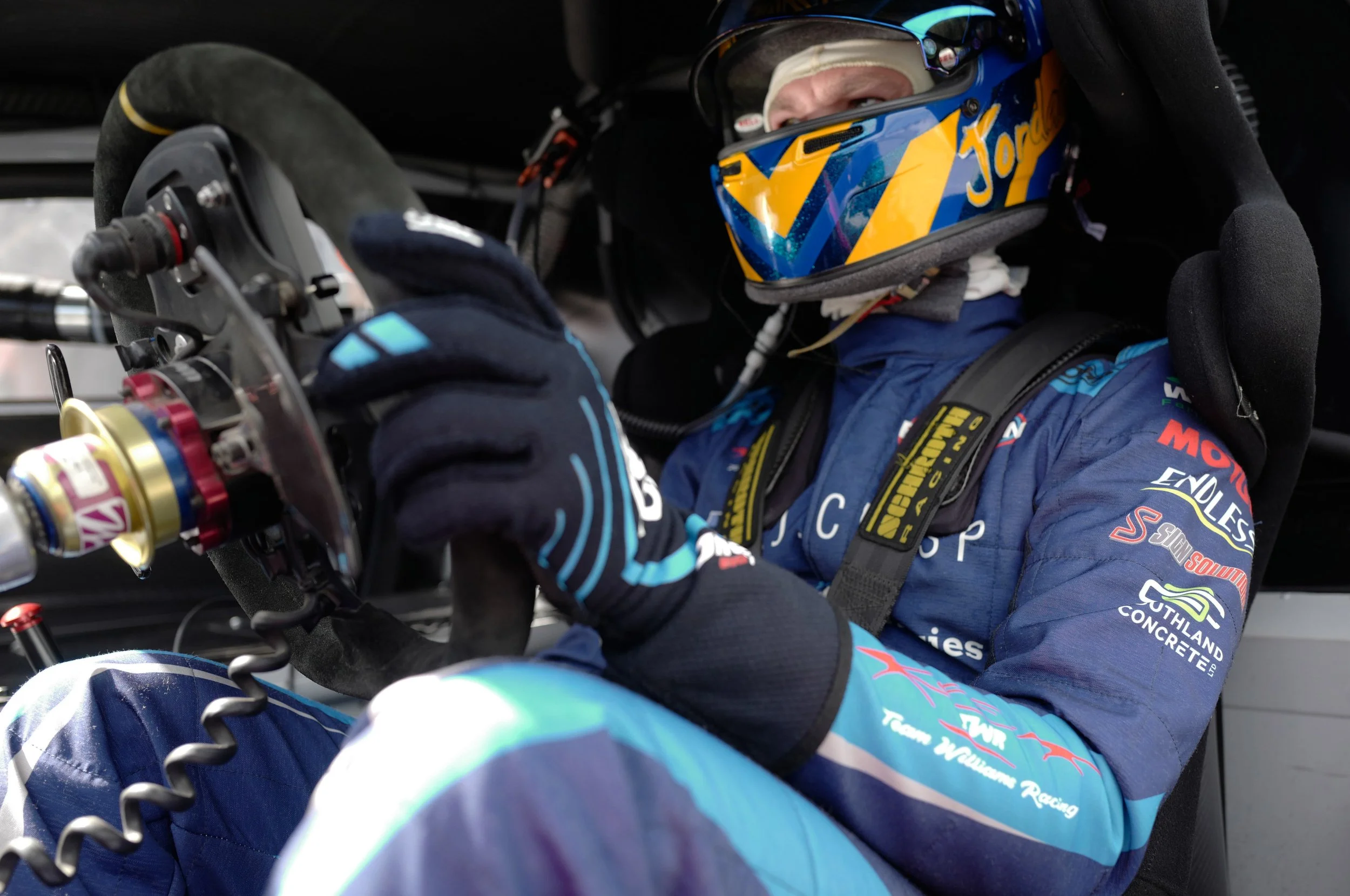 Close-up of a race car driver wearing a blue racing suit and helmet, sitting in a race car, gripping the steering wheel, with various sponsor logos on the suit.