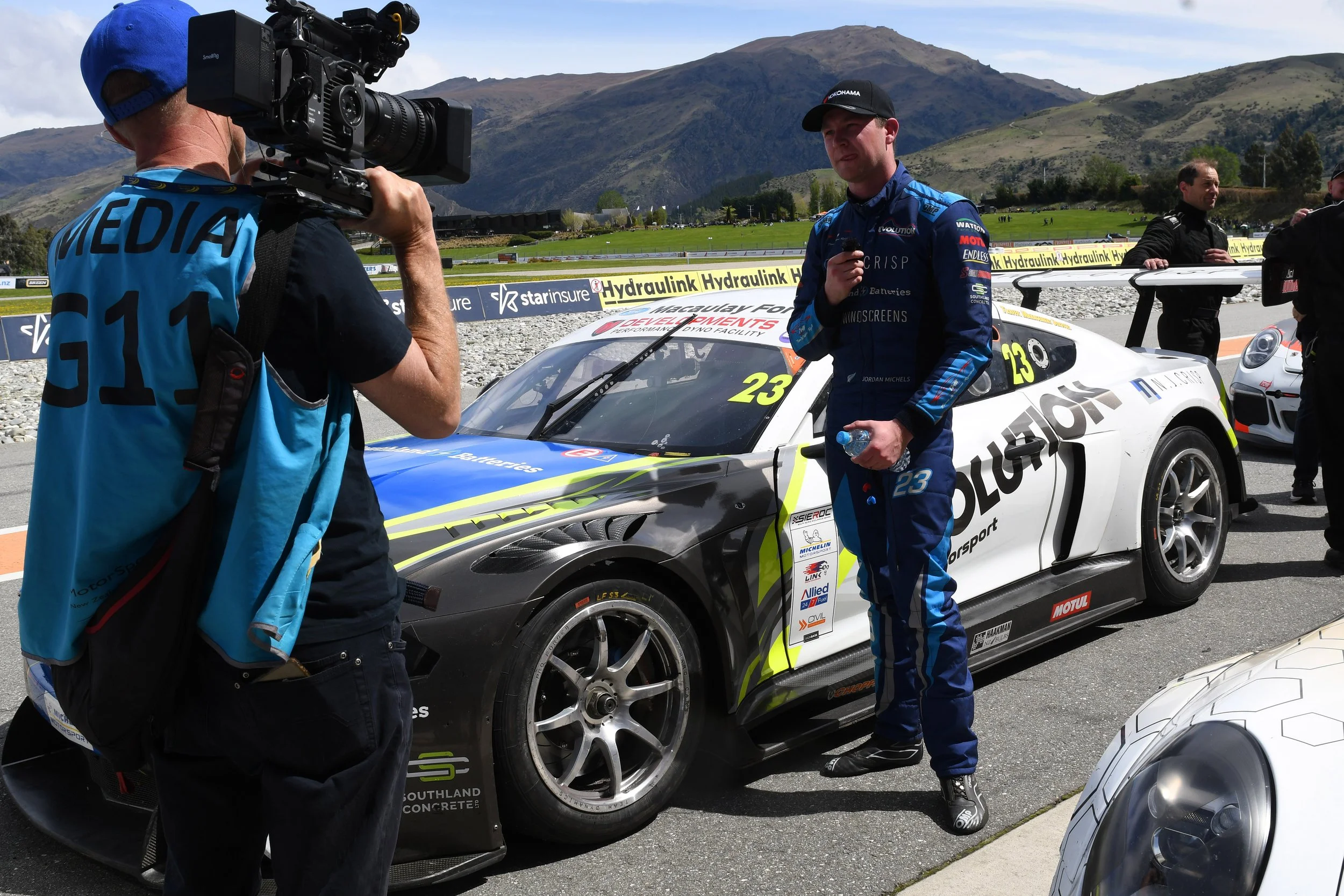 A race car driver in blue racing suit holding a water bottle, standing next to a white race car with yellow and black sponsor logos, being filmed by a cameraman with a professional video camera, in an outdoor setting with green mountains in the backg