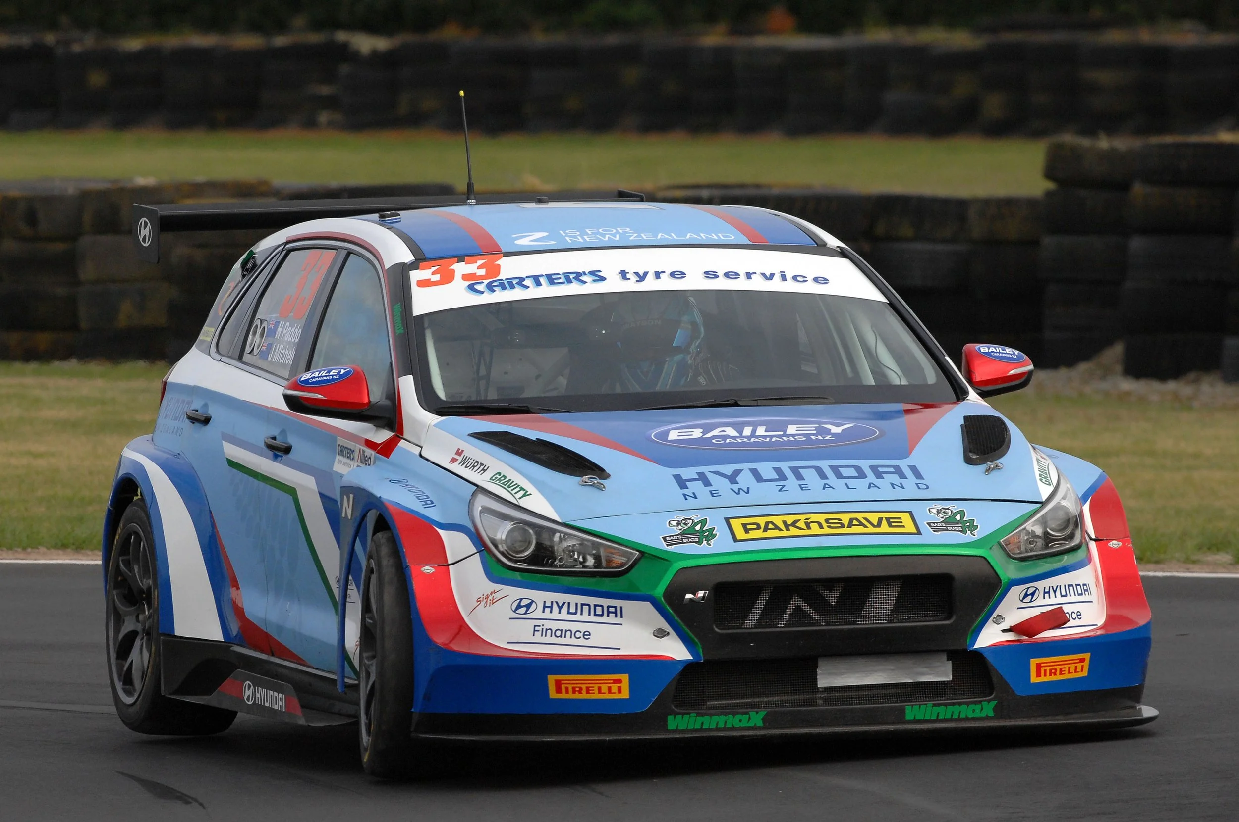 A blue, white, red, and green Hyundai rally car with various sponsor logos driving on a racetrack with tires stacked in the background.