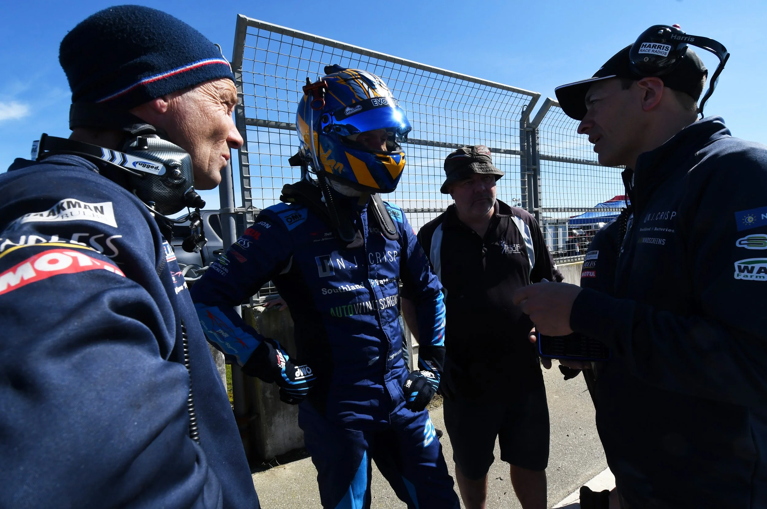 Group of race car drivers and team members standing near a fence at a race track on a sunny day, engaged in conversation.