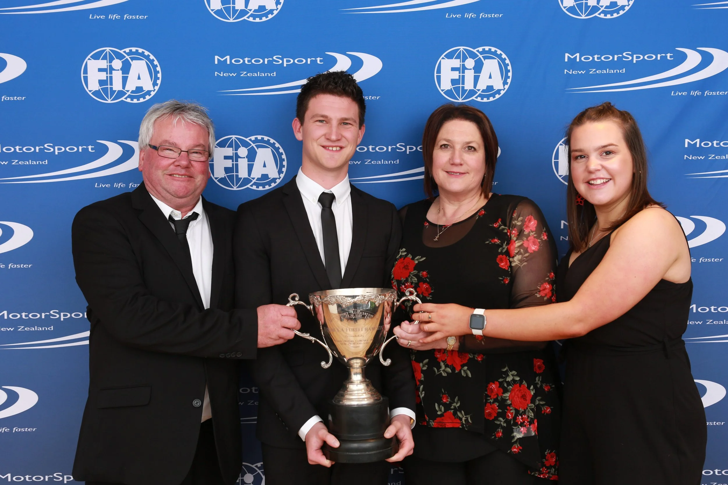 A group of four people, three women and one man, standing in front of a blue FIA MotorSport New Zealand backdrop. They are dressed formally and smiling, holding a silver trophy together.