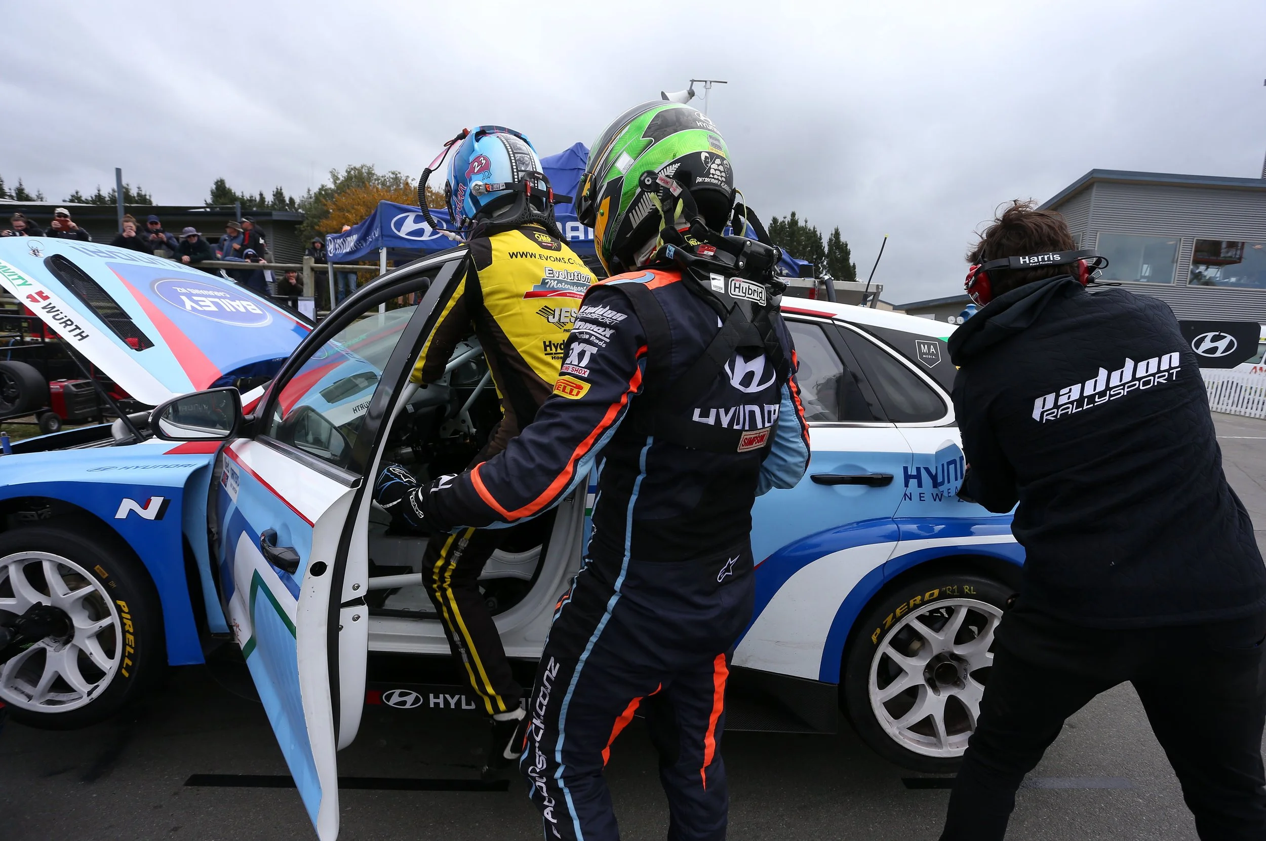 Two race car drivers in racing suits and helmets standing beside a Hyundai race car in a racing paddock area, with team members and spectators in the background.