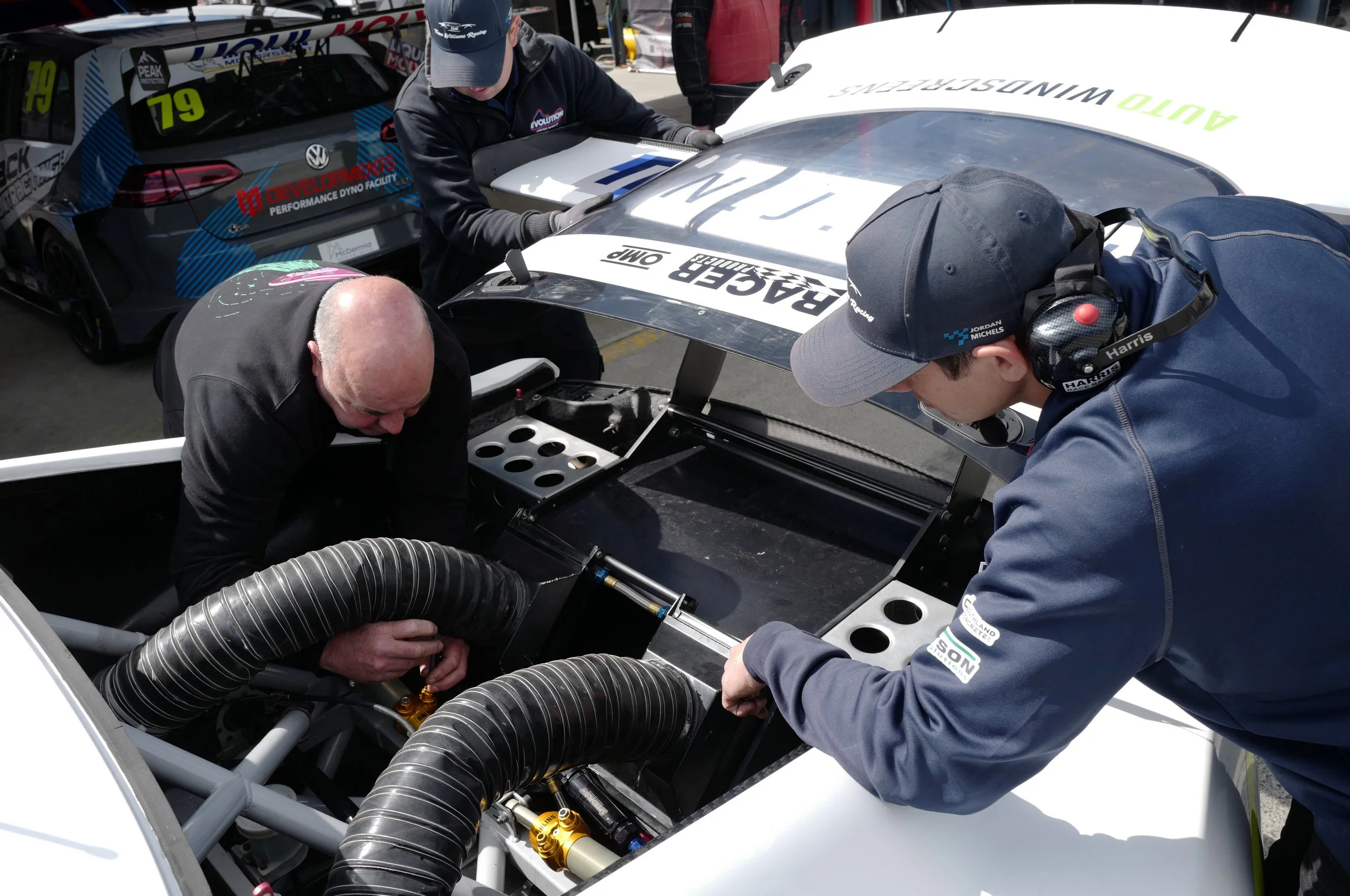 Three men working on a race car engine in a pit area. The car has a white body with sponsor logos, and a black race car with a blue and red design is visible in the background.