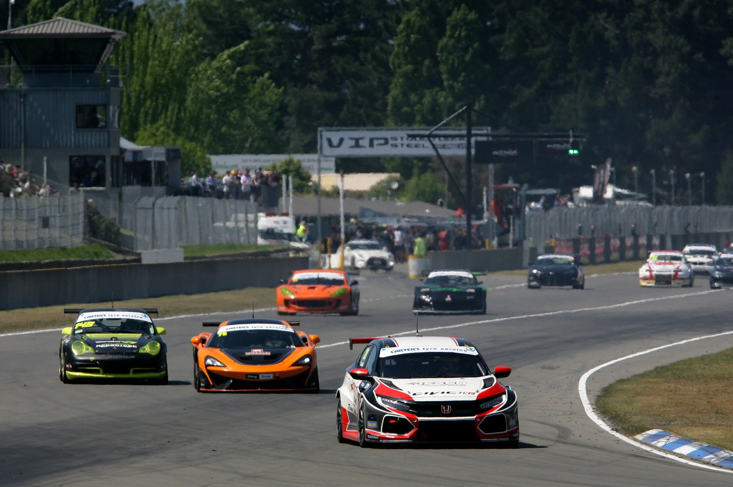 Race cars on a track with spectators and a tower in the background.