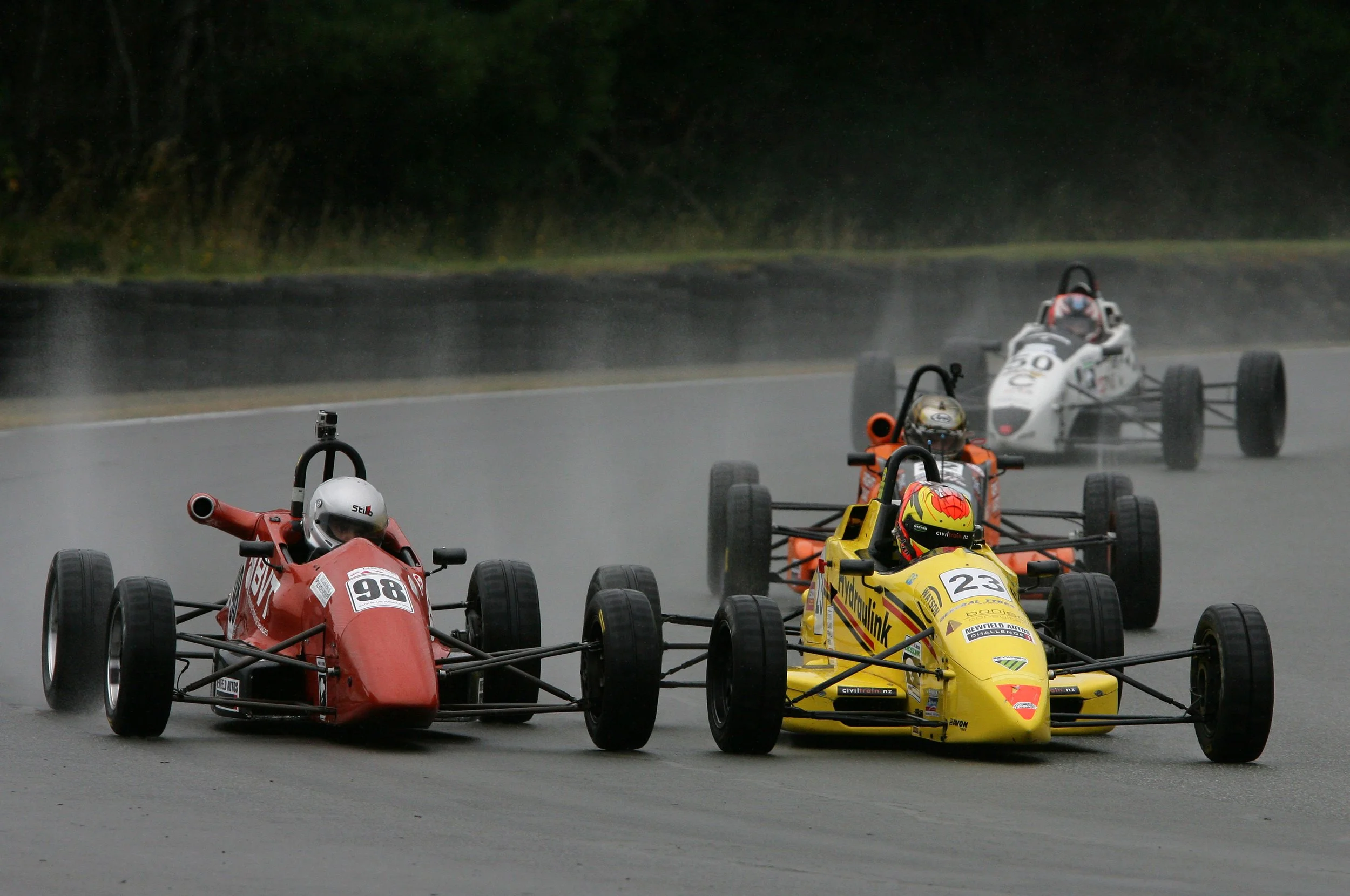 Race cars on wet track during a motorsport race, with drivers wearing helmets and racing suits.