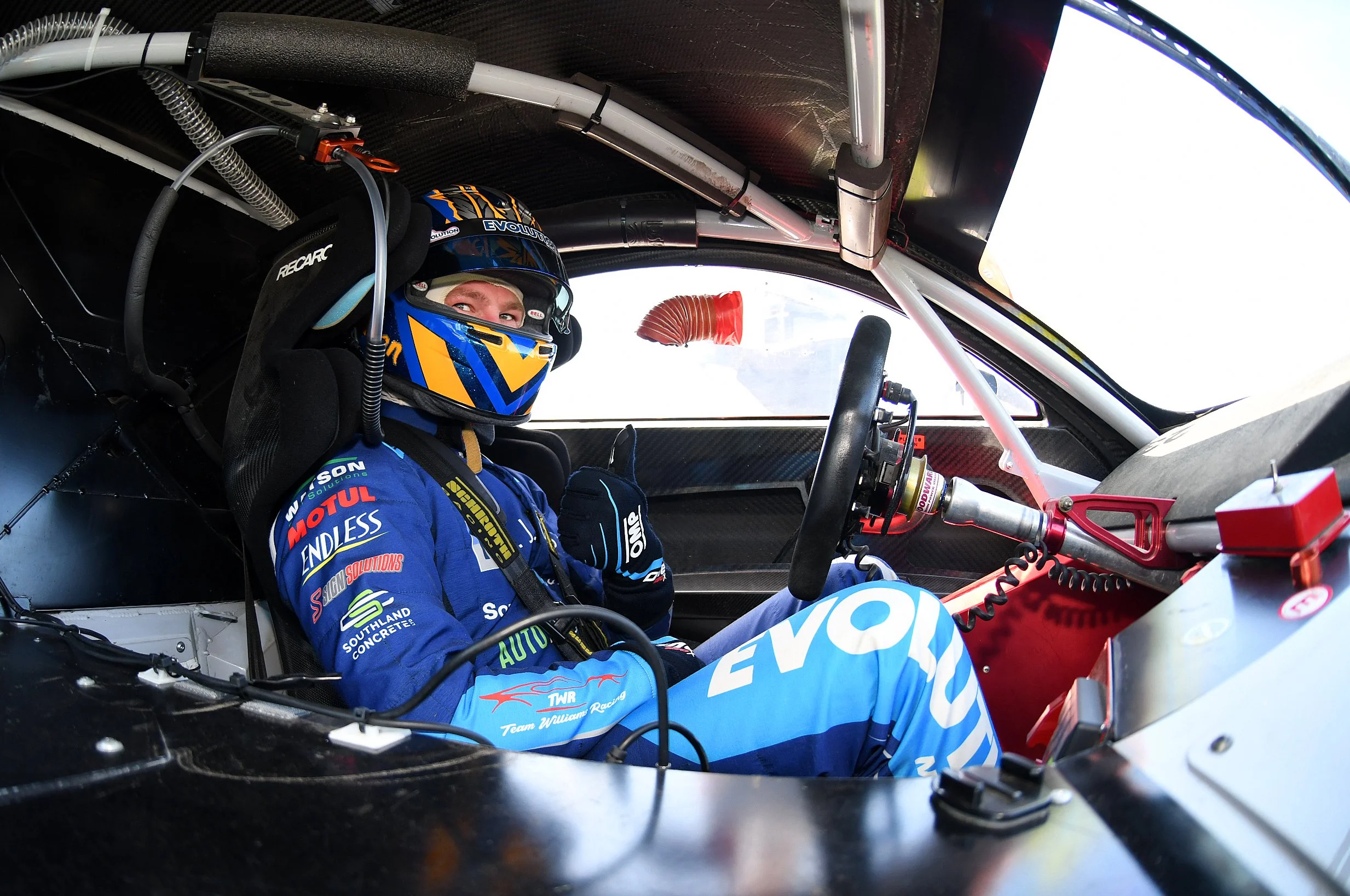 A race car driver wearing a blue racing suit and helmet sitting inside a race car cockpit.