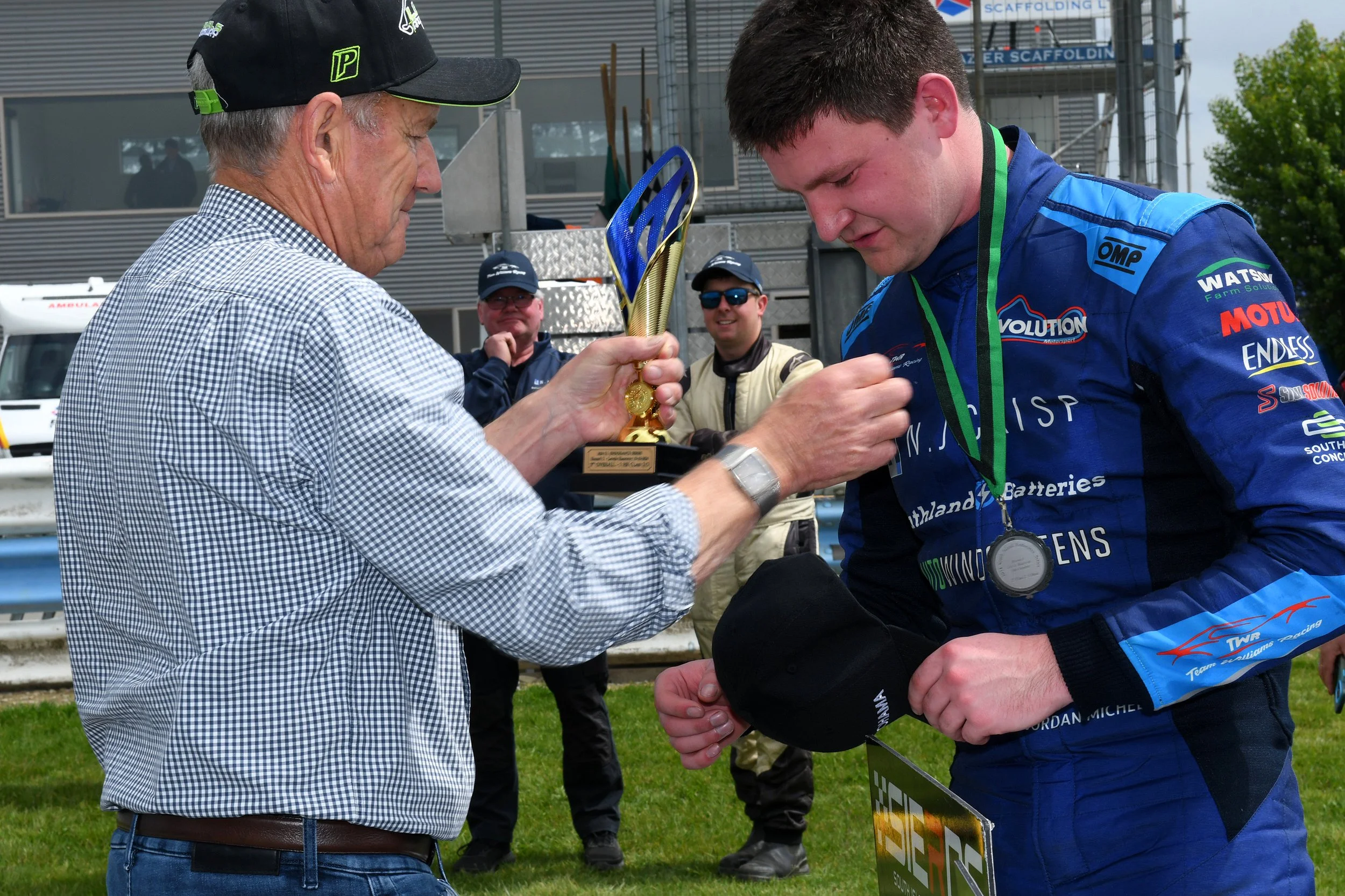 An older man in a checkered shirt and black cap awards a gold trophy to a young man in a blue racing suit, with two of the young man’s team members and a scaffolding structure visible in the background.