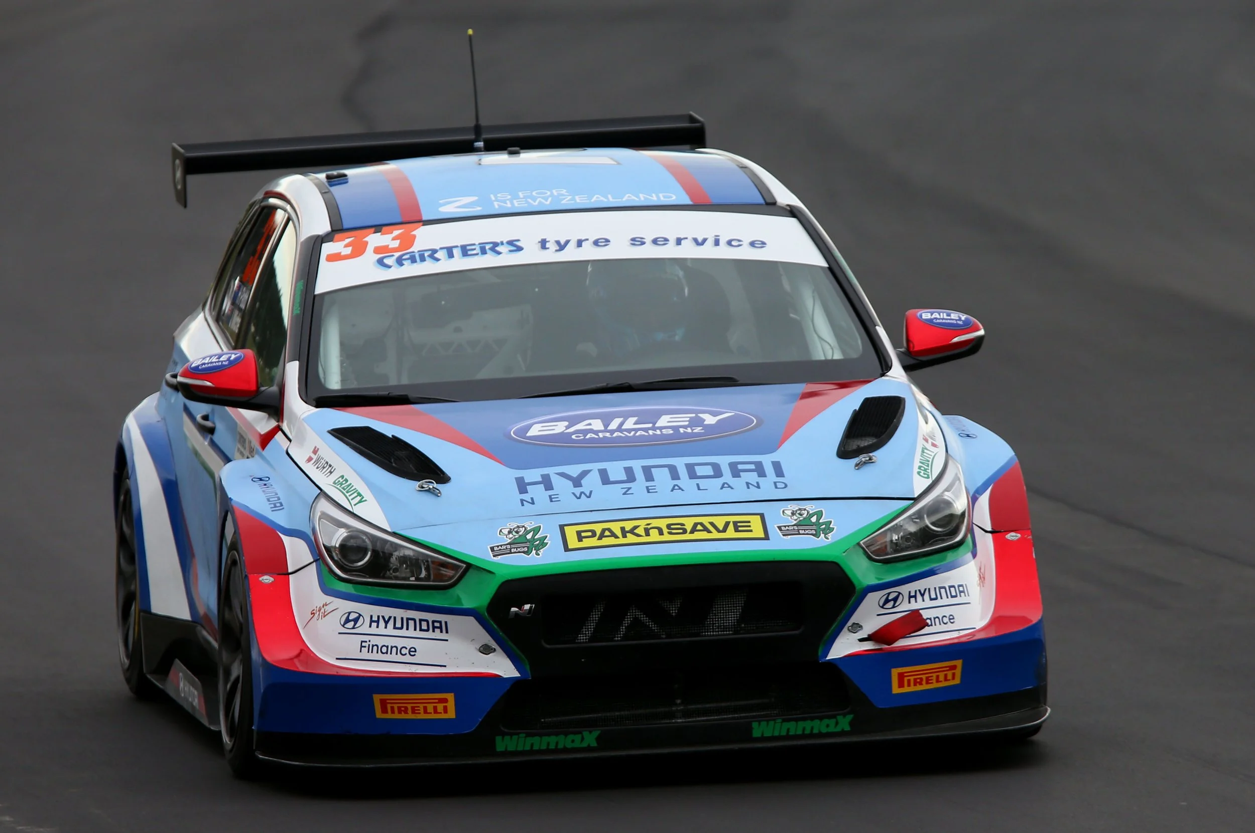 A blue, red, and white Hyundai race car on a racetrack, decorated with various sponsor logos including Pirelli, Hyundai, and sponsors from New Zealand, with a large rear wing and aerodynamic features.