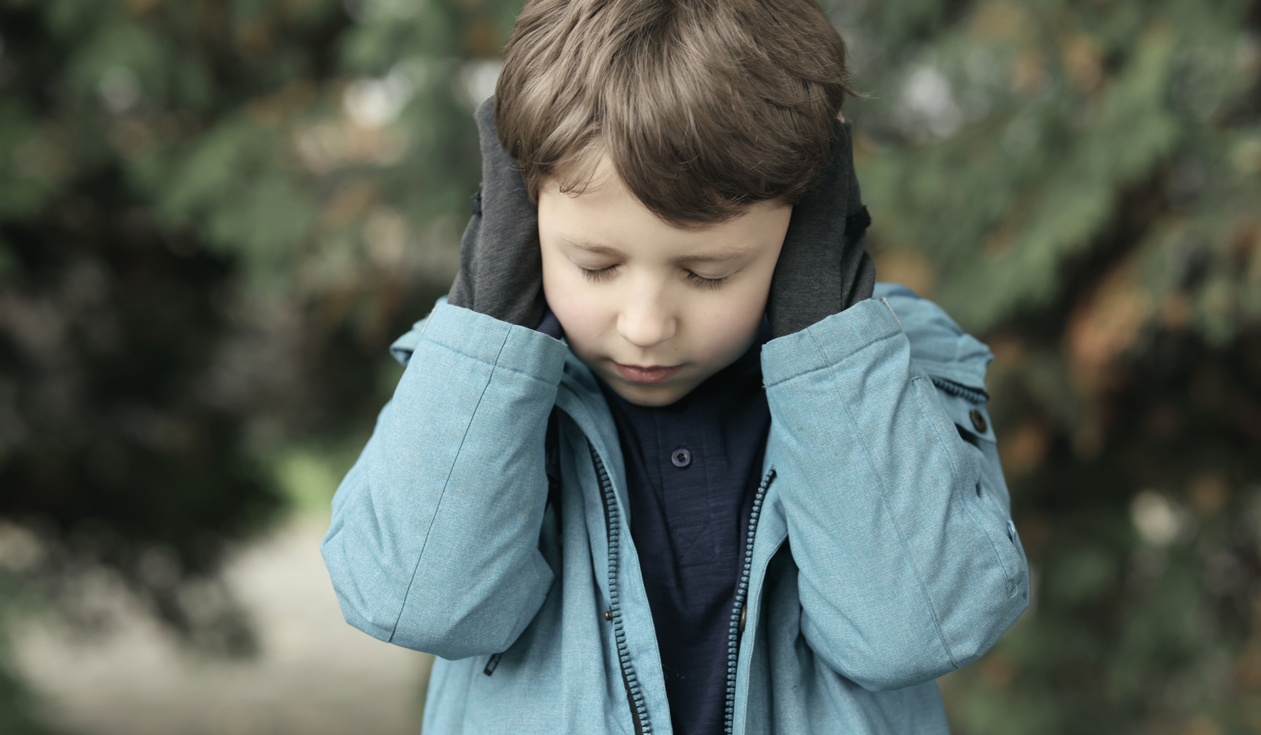 Child in a blue jacket covering his ears with eyes closed, a common sign of sensory overload