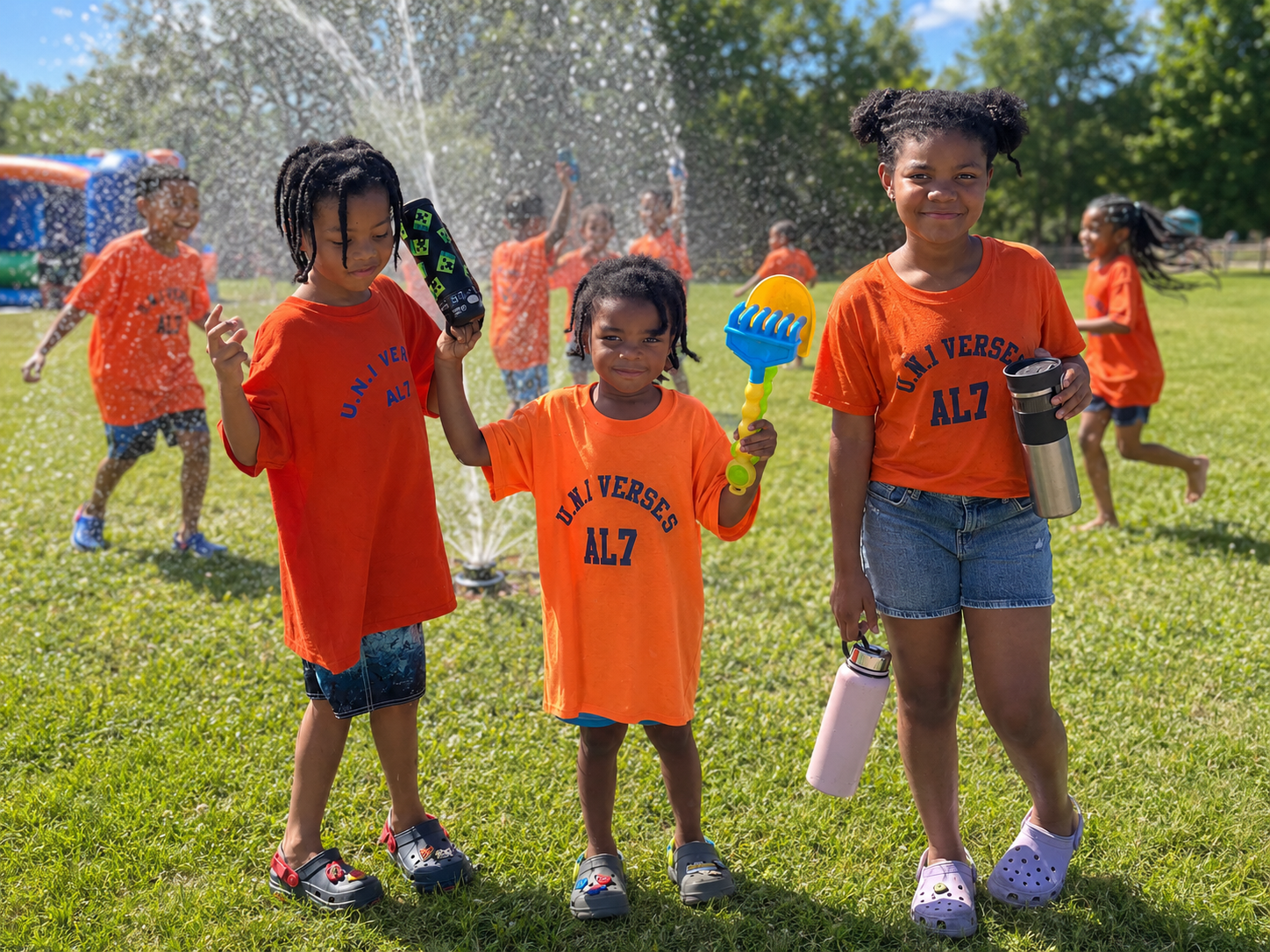 Children in orange shirts with 'U.N.I VERSes AL7' on front playing in a park on a sunny day, some holding water bottles and toys, others playing in the water spray, all with smiles.