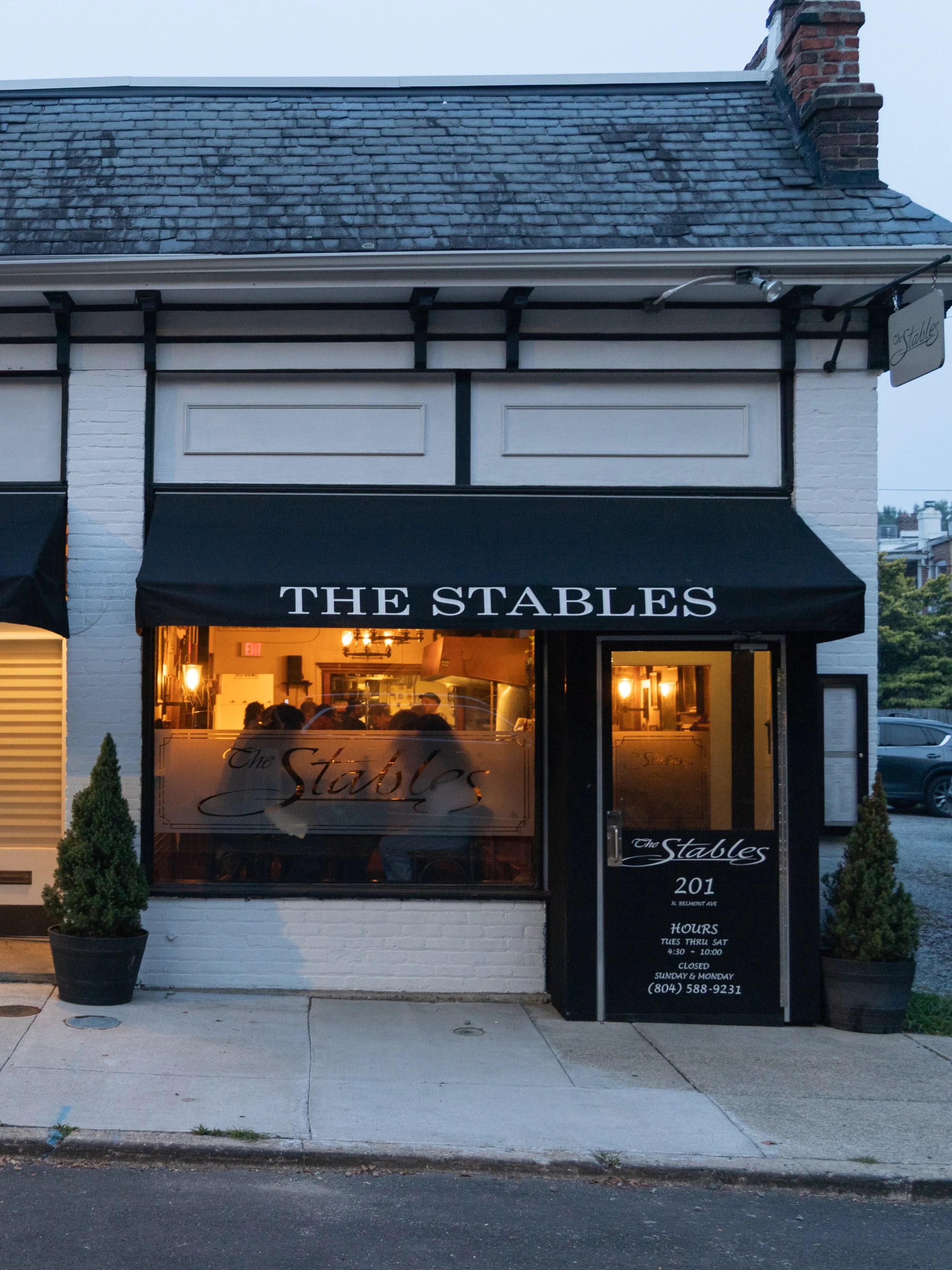 The exterior of a restaurant called The Stables with a black awning and large front window showing an interior with people dining. The building has a white brick facade, a slate roof, and a brick chimney. There are two small potted trees on either side of the entrance, and a sidewalk in front.