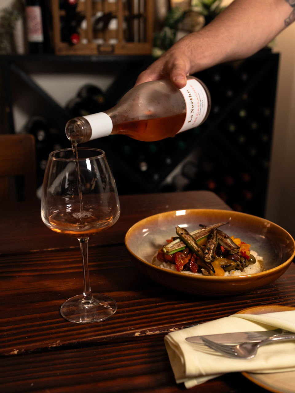 Person pouring rosé wine into a glass over a wooden table, with a bowl of mixed grilled vegetables and rice nearby, in a cozy dining setting.