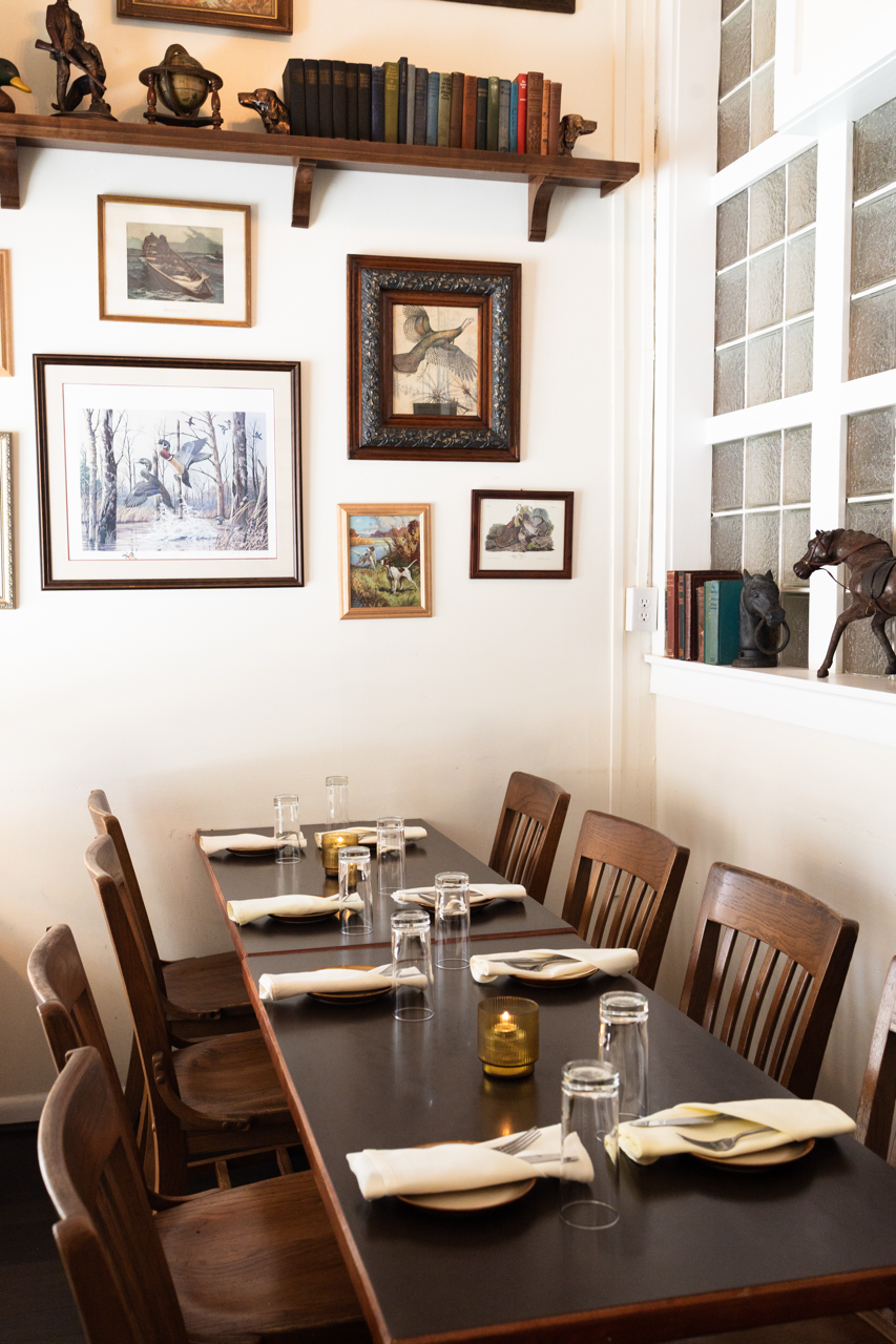A dining room with a long dark wooden table set for six, featuring white napkins, glasses, and small candles. The white wall behind is decorated with multiple framed artwork and photographs, and a wooden shelf holding a stack of books and decorative items. To the right, a window with textured glass blocks allows natural light into the room.