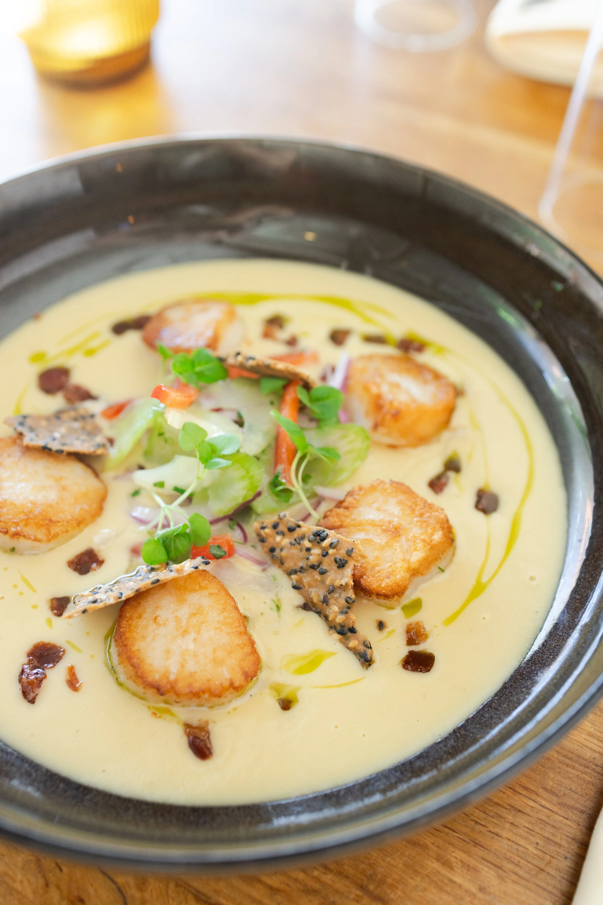 Close-up of a bowl of creamy soup topped with microgreens, crispy fish skin, croutons, and a drizzle of oil, served on a wooden table.