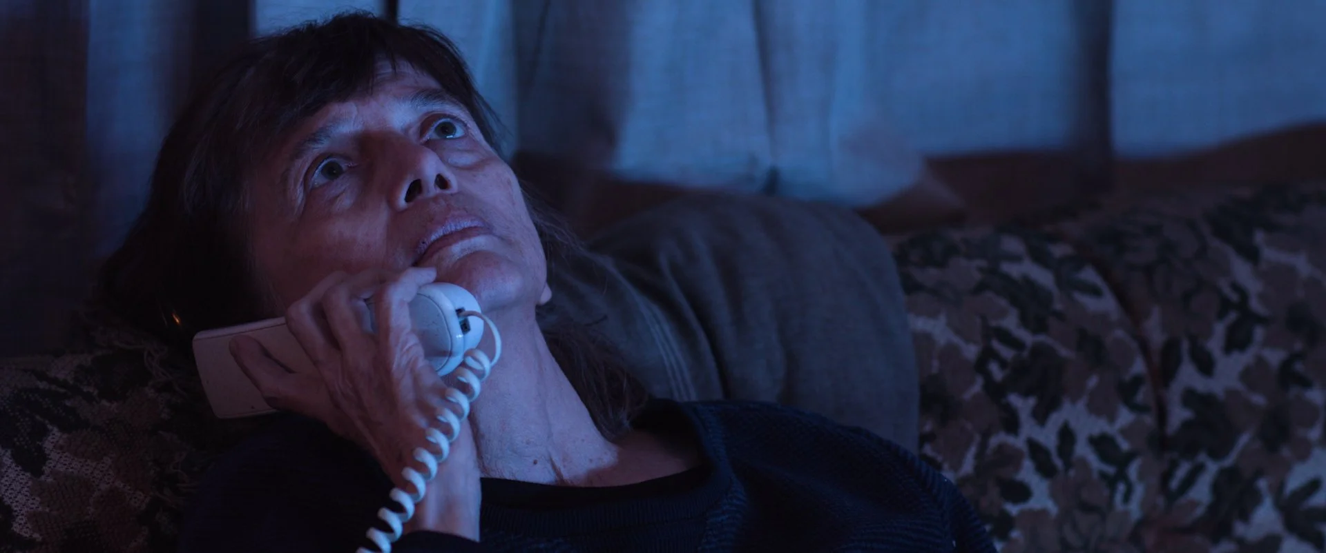 A woman with a distressed expression lying on a couch, holding a landline phone receiver to her ear, illuminated by blue light.