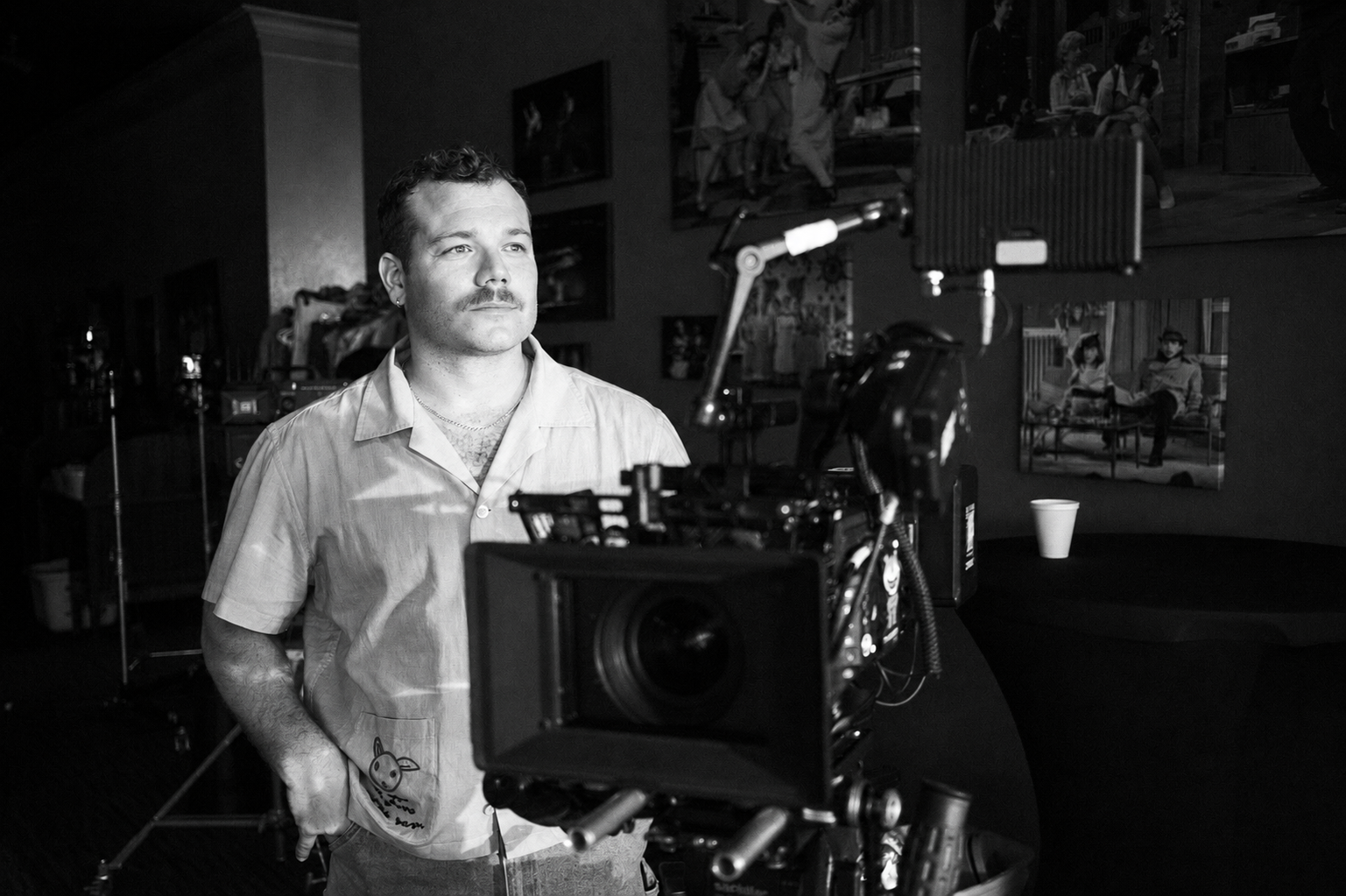 A man stands behind a professional camera on a film set, looking off into the distance. The scene is in black and white, with photographs hanging on the wall in the background.