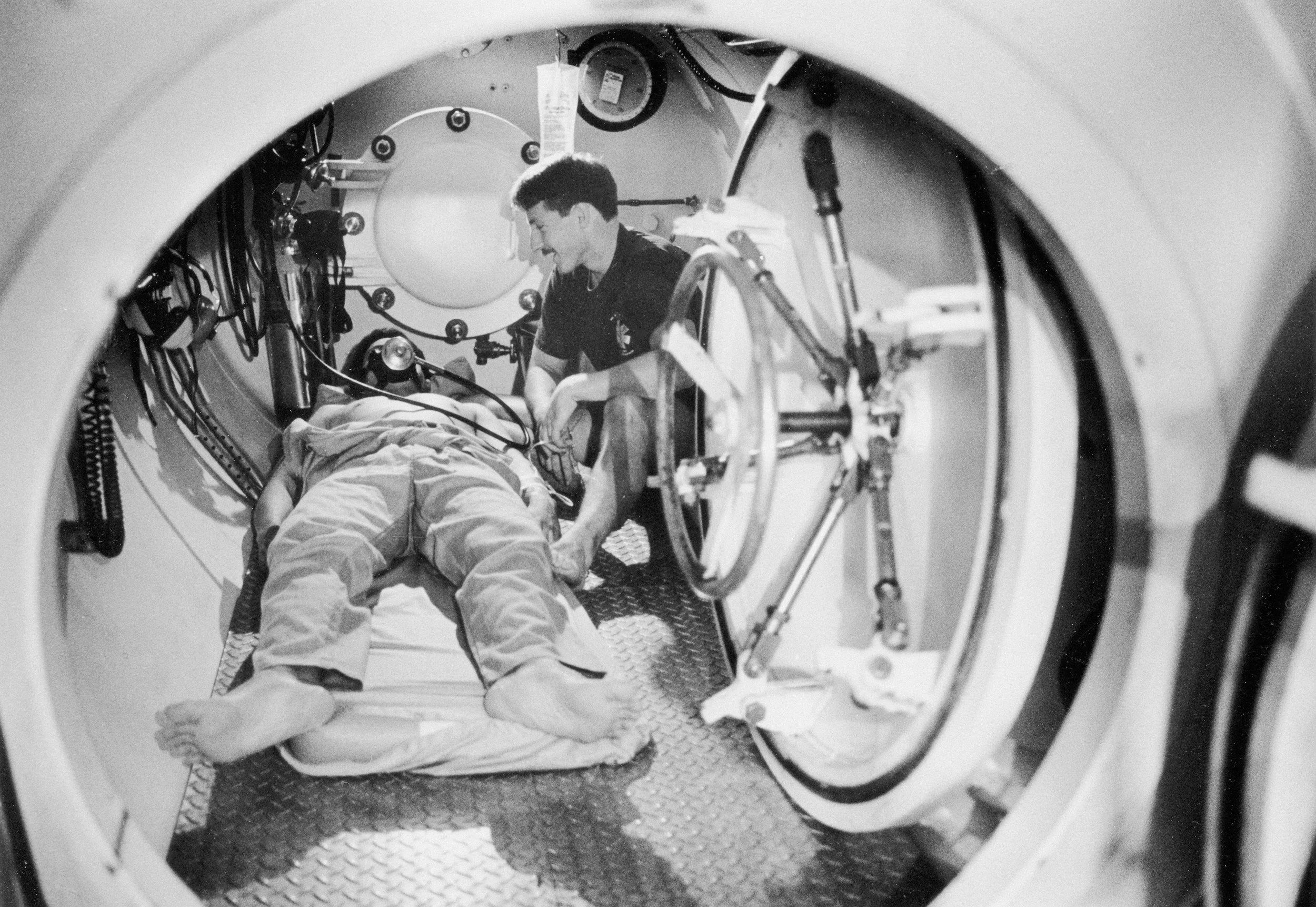 A person lying on a hospital bed or gurney inside a medical imaging machine, viewed through the circular opening of the machine.