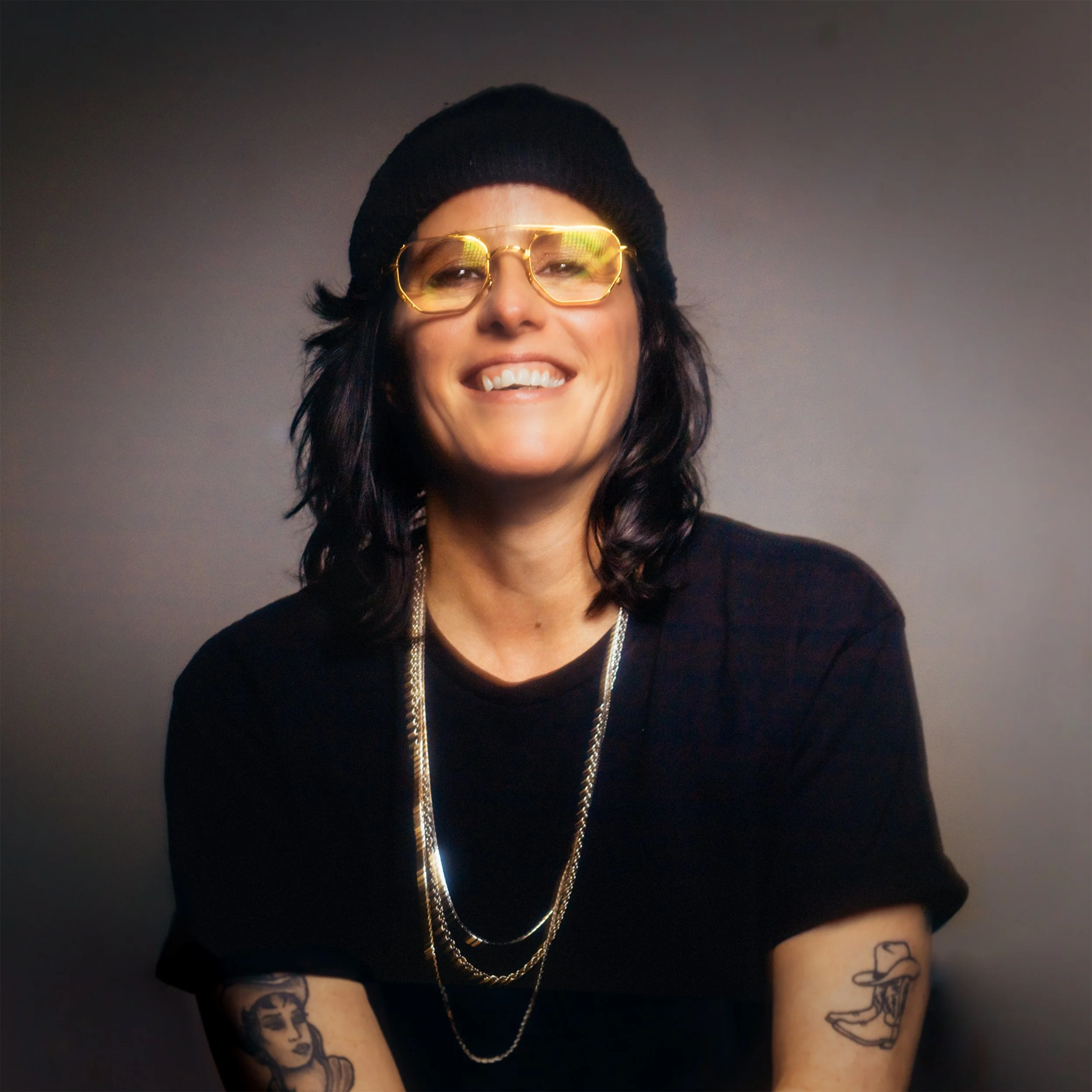 A person with black hair, wearing gold-rimmed glasses, a black beanie, and a black shirt, smiling, with visible tattoos on their arms, against a plain background.