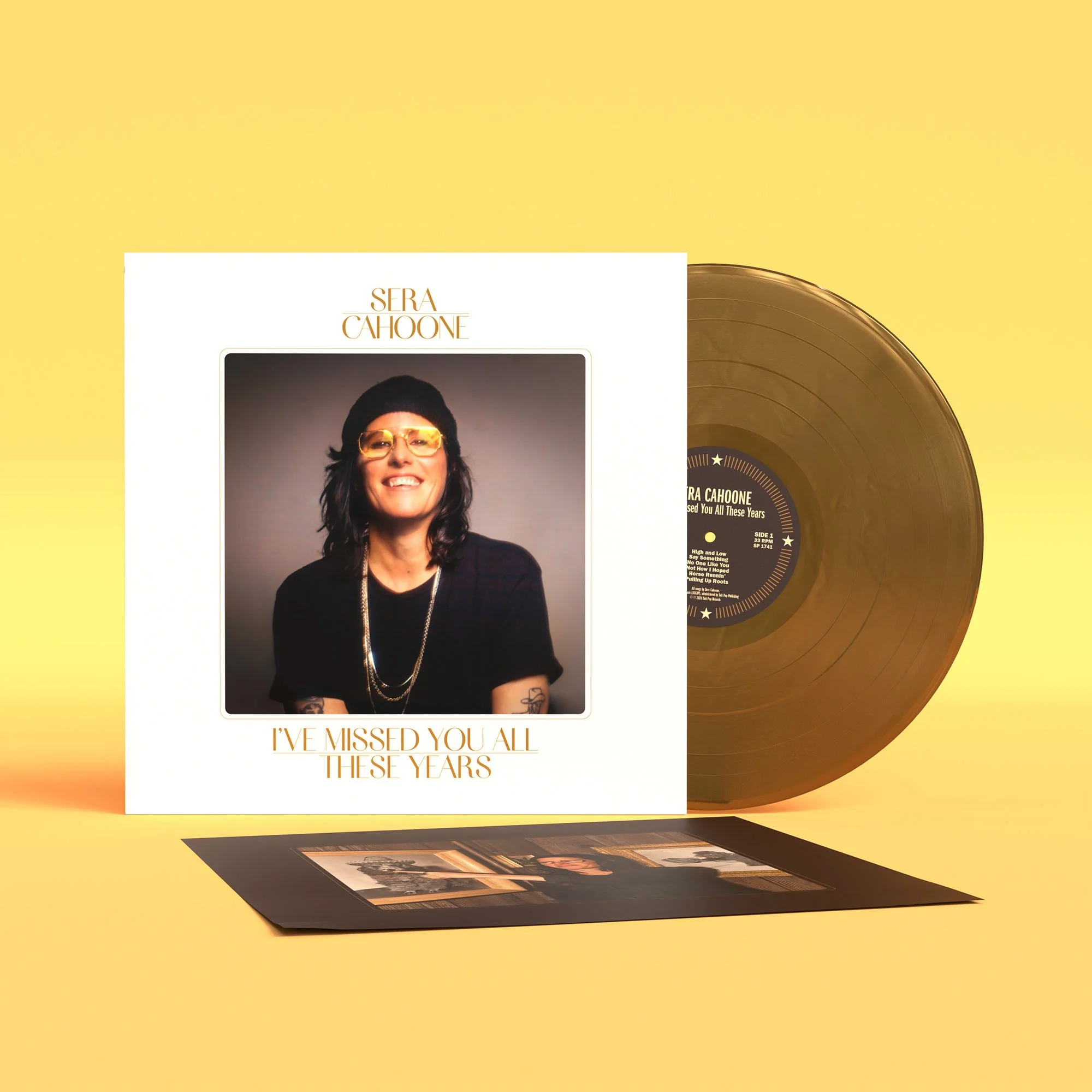 A gold vinyl record is partly out of an album sleeve that features a photograph of a smiling woman with dark hair and gold-framed glasses, all set against a bright yellow background.