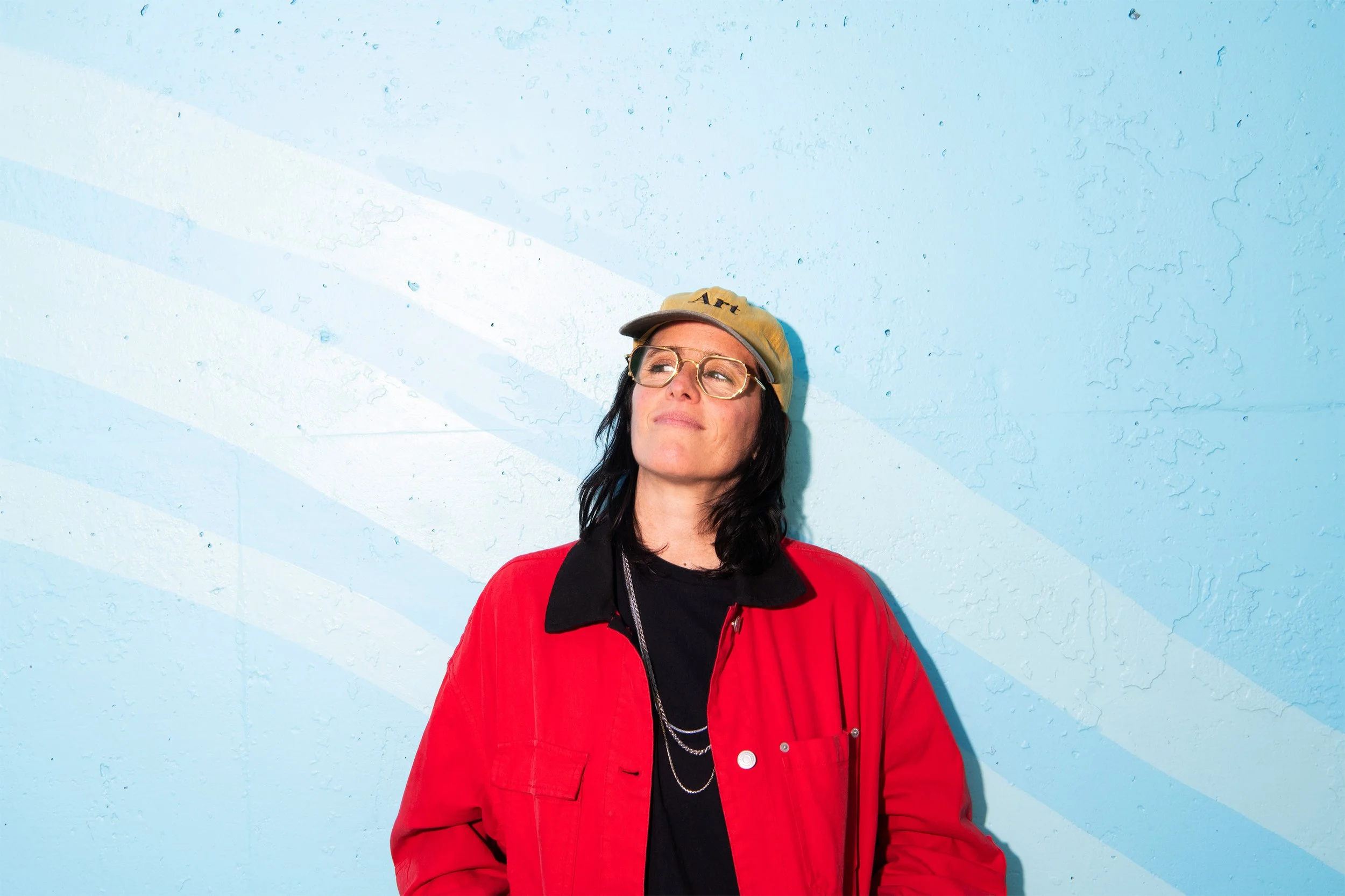 A woman in a red jacket standing against a light blue wall with diagonal white stripes. She is wearing glasses, a beige cap with the word 'Art' on it, and has dark hair.