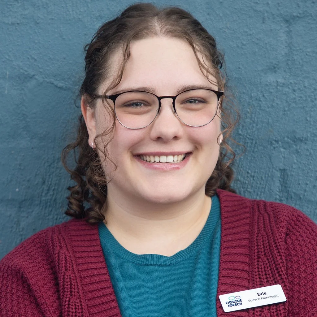 Young woman with glasses smiling against a blue wall, wearing a teal shirt and maroon cardigan, with a name tag reading 'Evie, Speech Pathologist'.