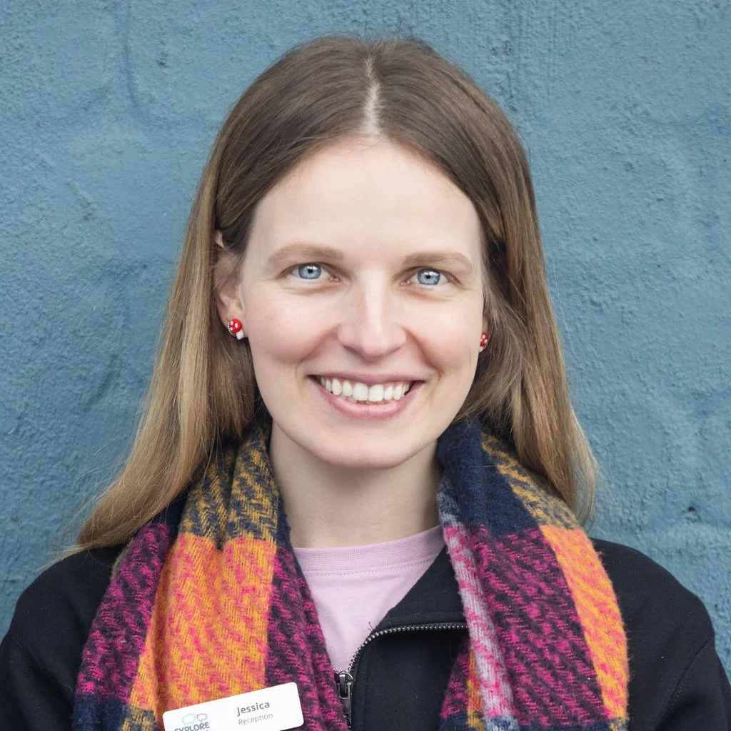Headshot of a young woman with long light brown hair, bright blue eyes, smiling, wearing a pink shirt, a colorful plaid scarf, a black jacket, red flower earrings, and standing against a blue textured wall.
