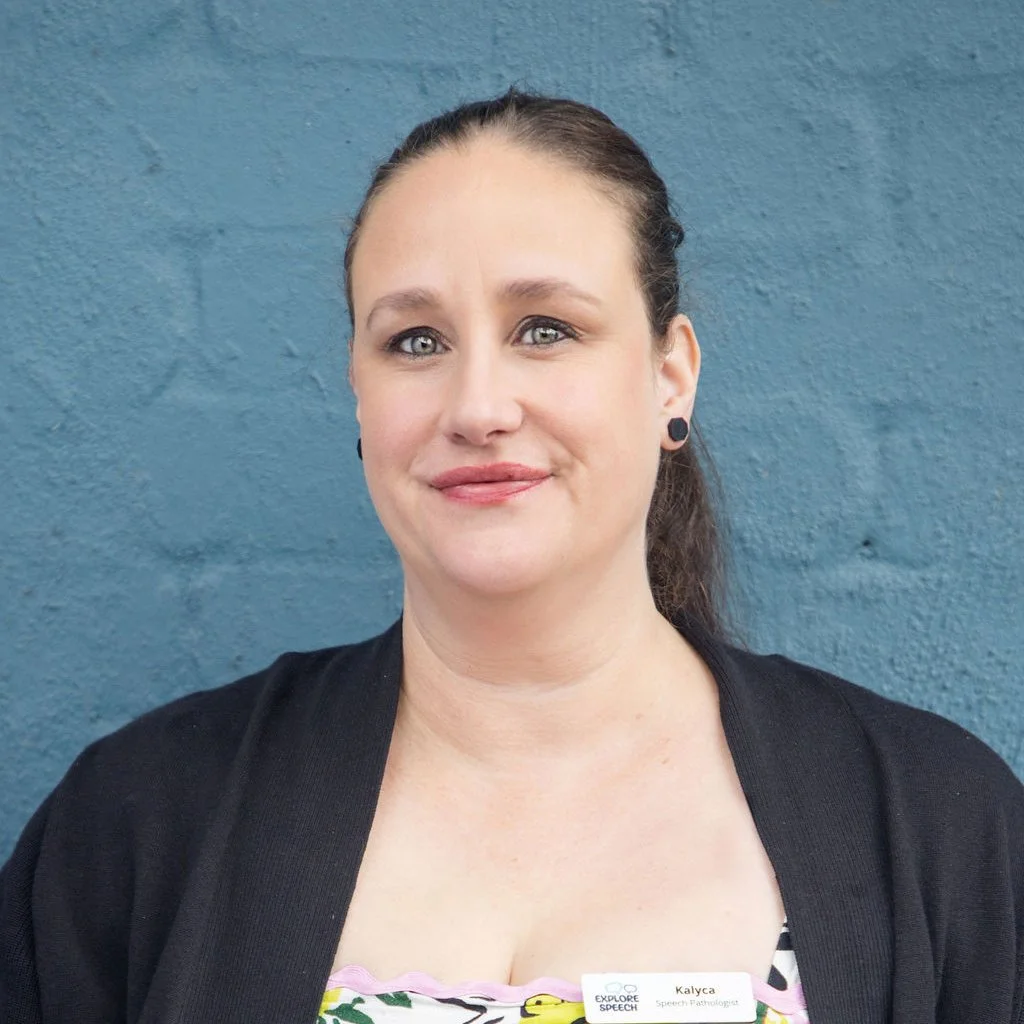 A woman with dark hair in a ponytail, wearing black earrings and a black cardigan, standing in front of a blue wall.