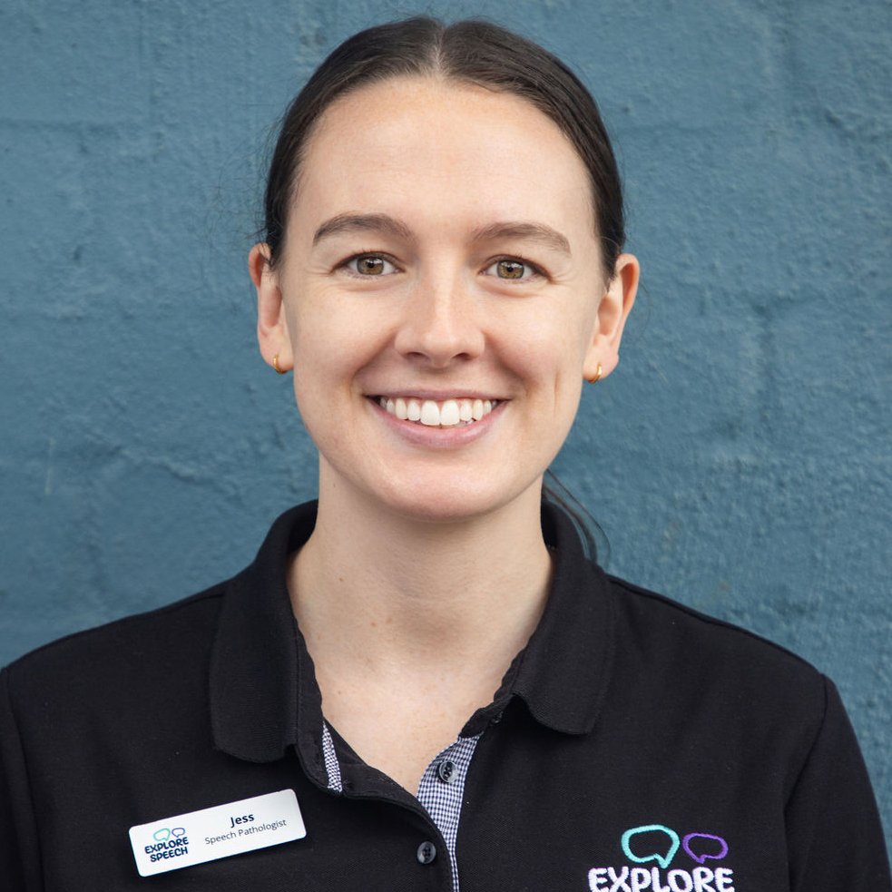 A smiling woman with dark brown hair wearing a black polo shirt with a name tag that says Jess, and a logo that says Explore Speech, standing in front of a textured blue wall.