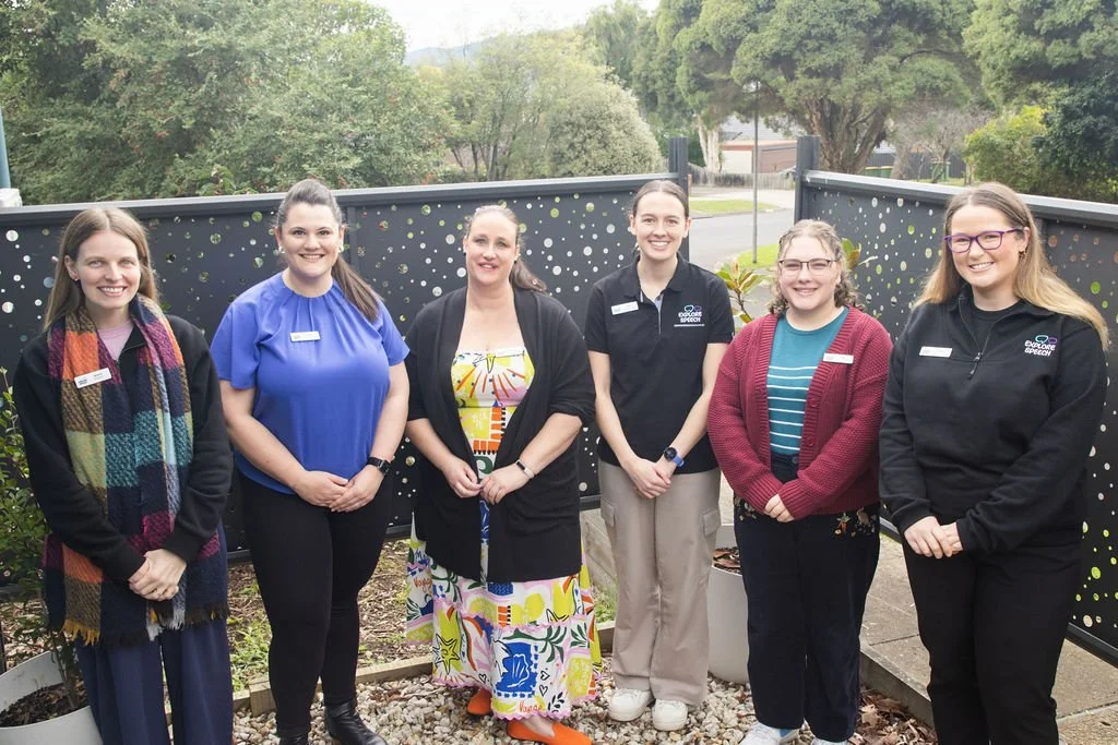 Six women standing outdoors on a balcony, smiling at the camera. Explore Speech staff.