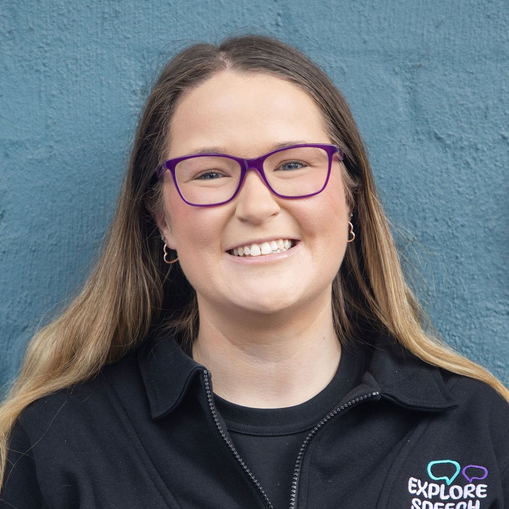 A young woman with long brown hair, purple glasses, and hoop earrings, smiling and wearing a black jacket with a logo that says 'Explore speech' standing in front of a blue wall.
