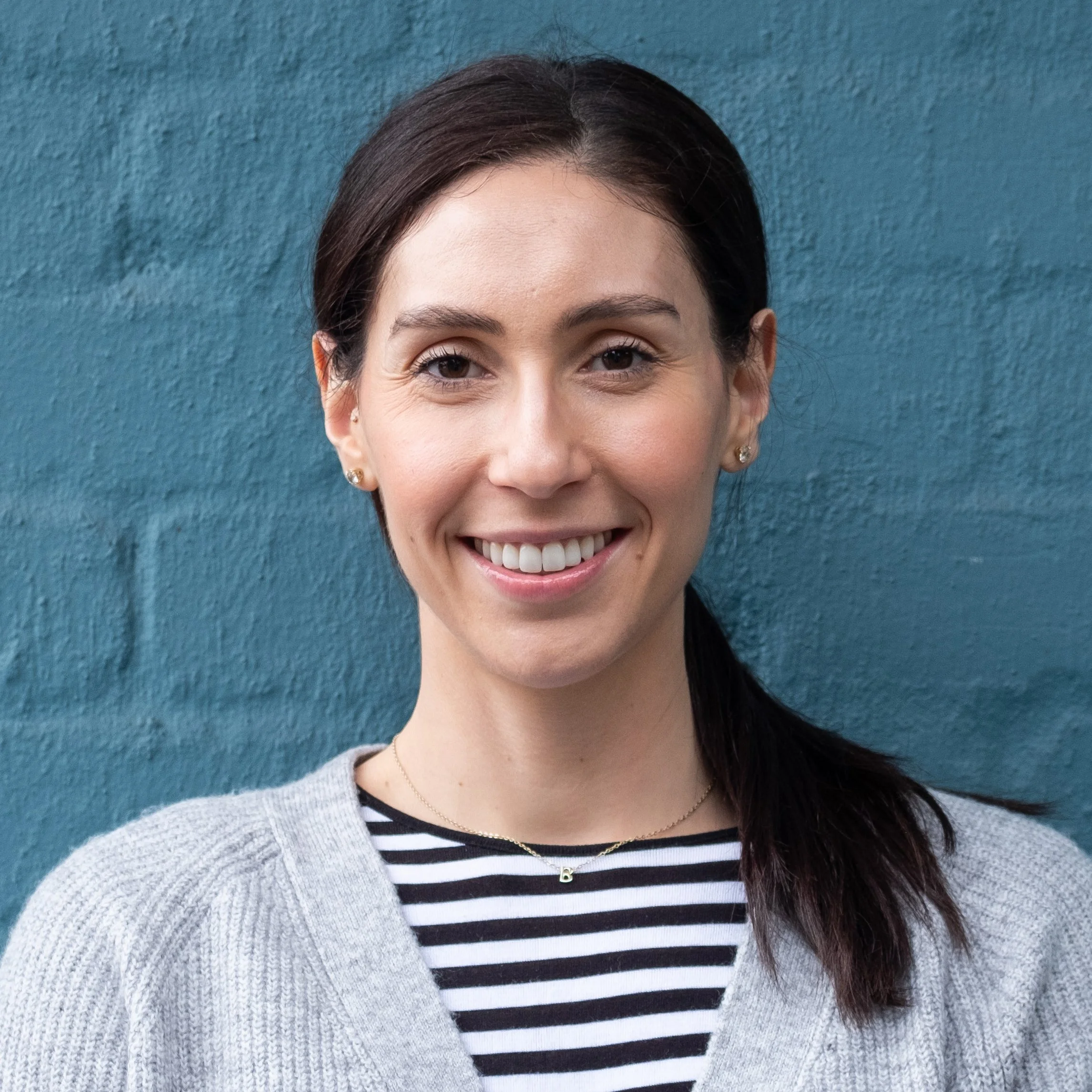 Smiling woman with dark hair in a ponytail, wearing earrings, a striped shirt, a necklace with a 'B' initial, and a gray knit cardigan, standing in front of a teal textured wall.