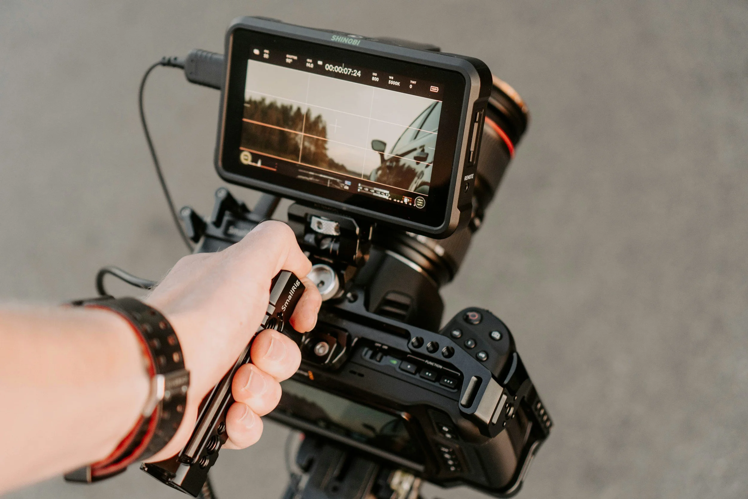 Close-up of a video camera monitor displaying a scene with trees and a vehicle, with a hand adjusting the camera settings.