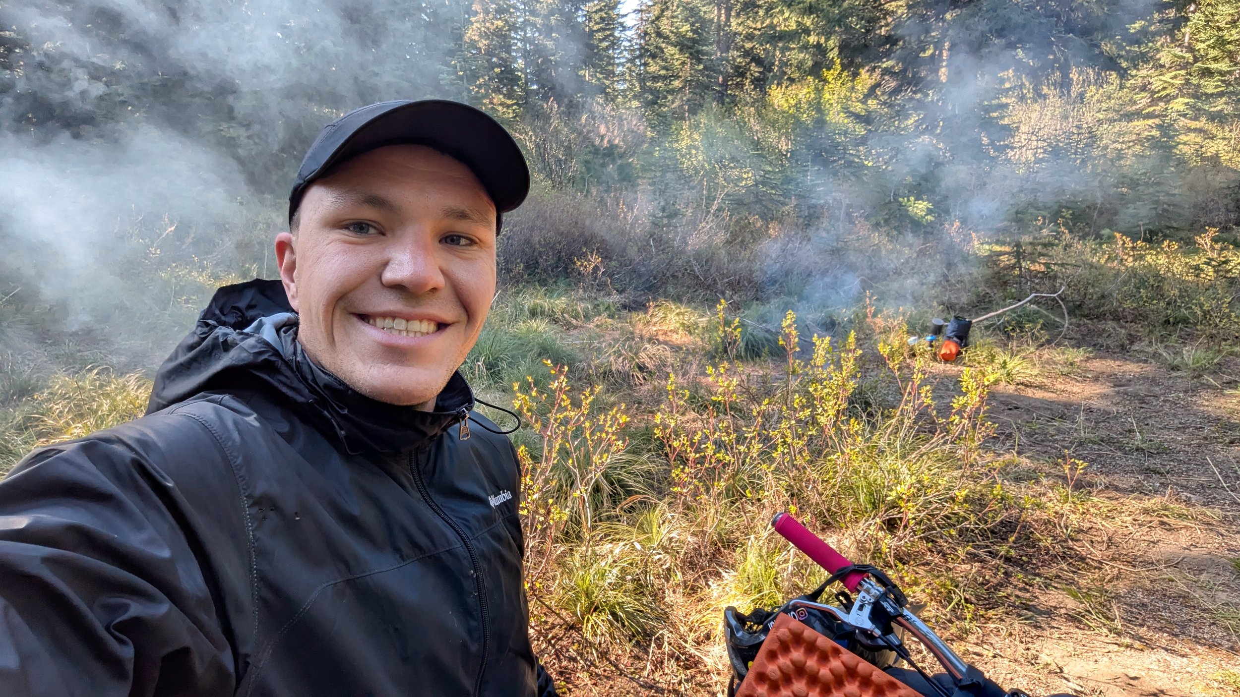 A smiling man in a black jacket and cap takes a selfie in a forested area with a smoky background. He is holding a bicycle with a pink handlebar, and the forest includes tall trees and green foliage.
