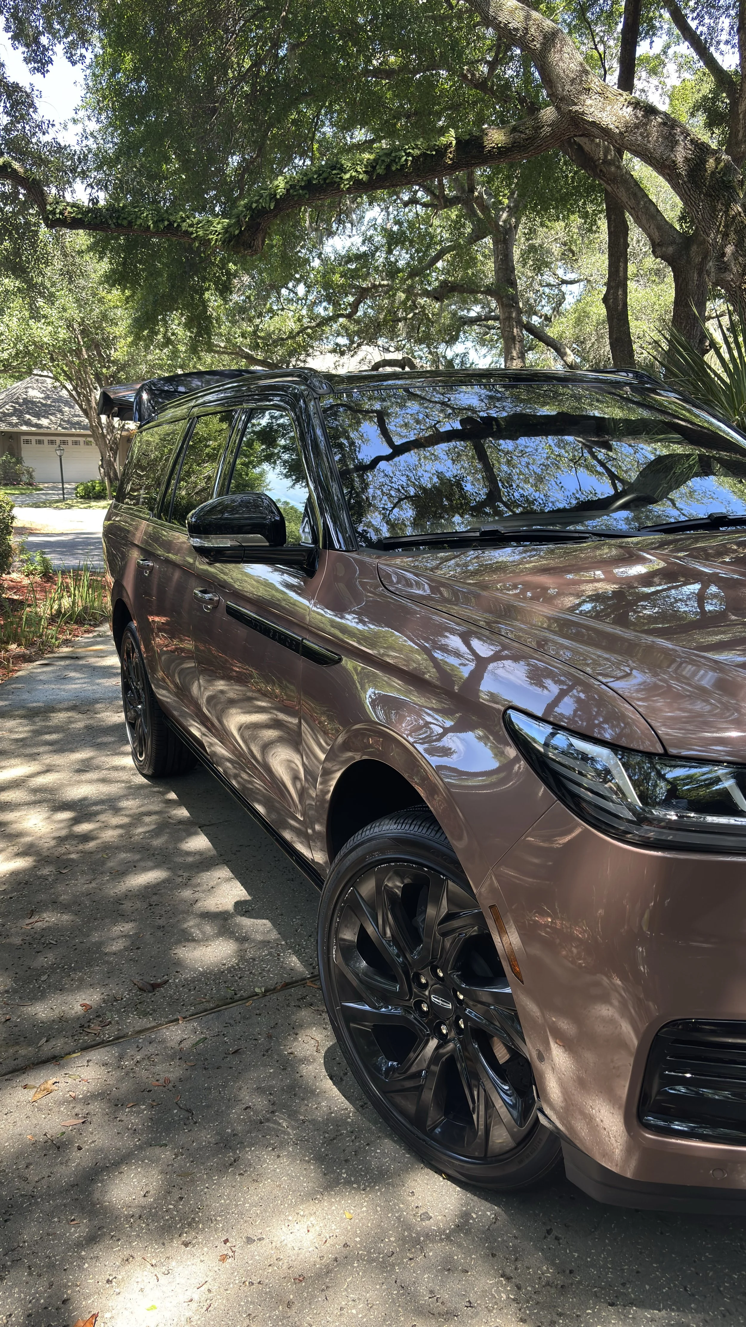 A brown SUV parked on a driveway under a tree with green leaves, with reflections of the tree in the car's windows and body.