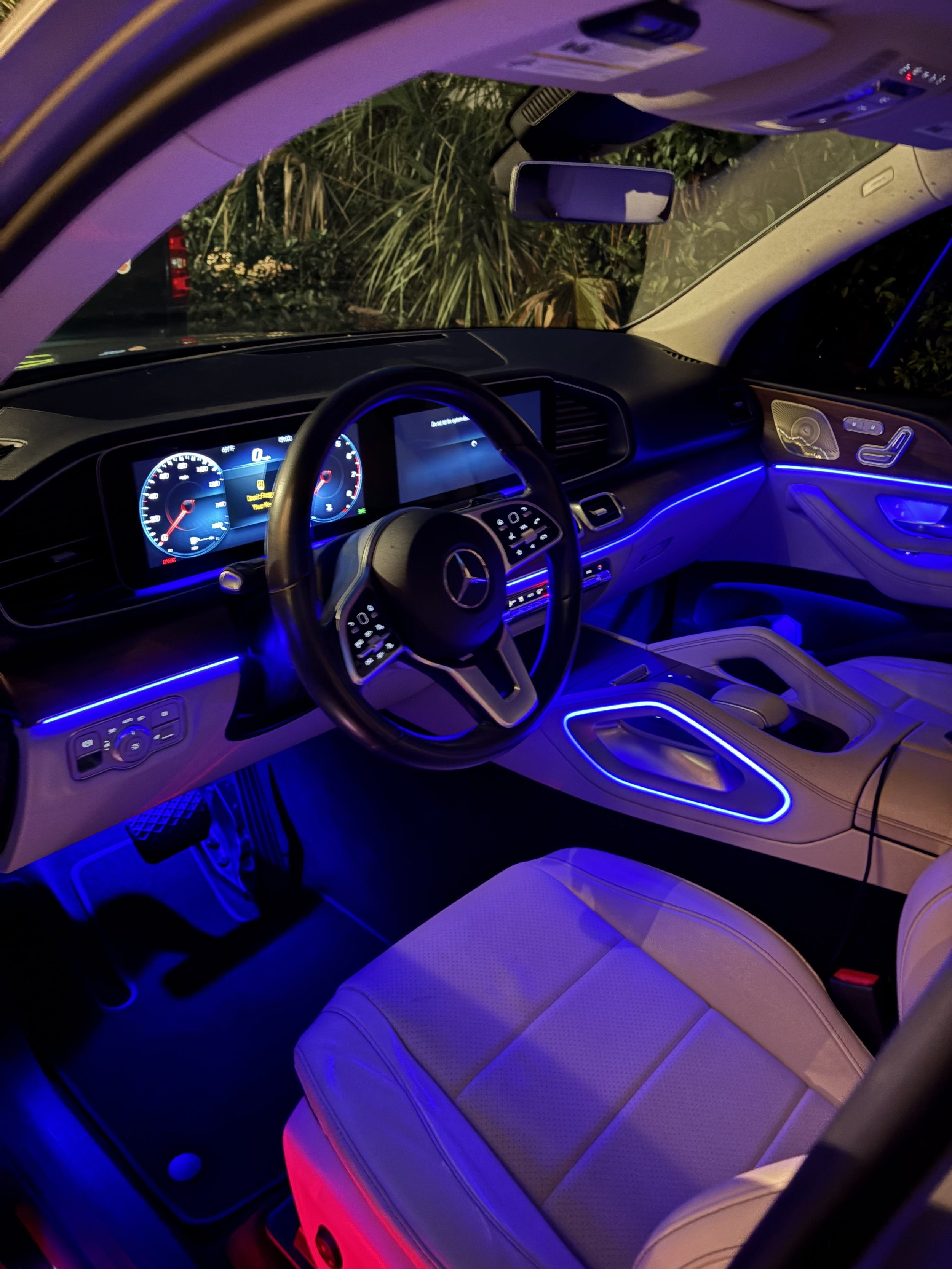 The image shows the interior of a luxury car illuminated with blue and red ambient lighting. The dashboard features digital displays, and the seats are made of light-colored leather with contrasting stitching.