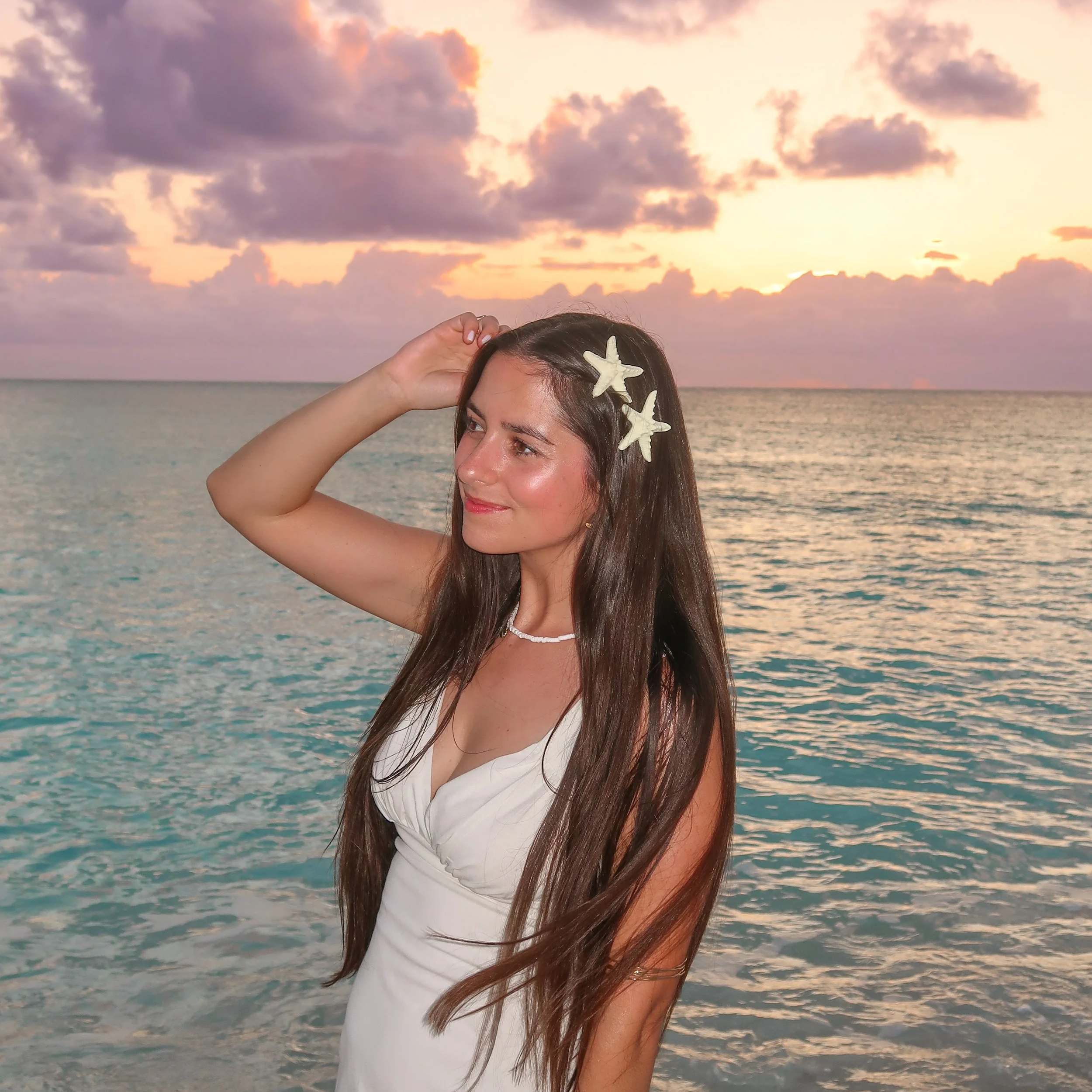 A woman with long dark hair and starfish hair clips stands at the beach during sunset, with the ocean and colorful sky in the background.