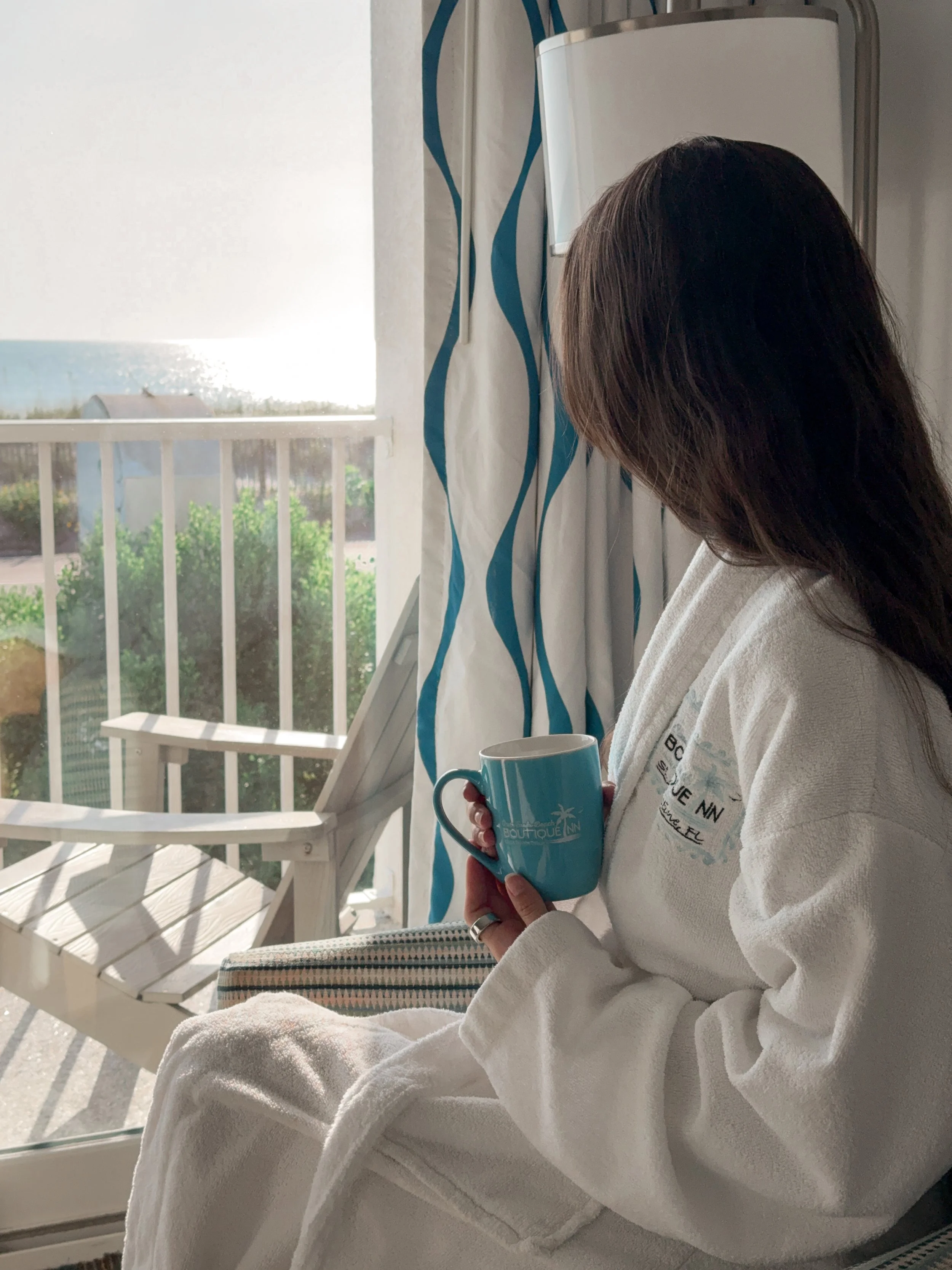 A woman with brown hair, seen from the side, is sitting by a window wearing a white bathrobe and holding a blue mug. The window shows an outdoor porch with a white railing and a bench, and beyond it, a view of a body of water with a distant shoreline