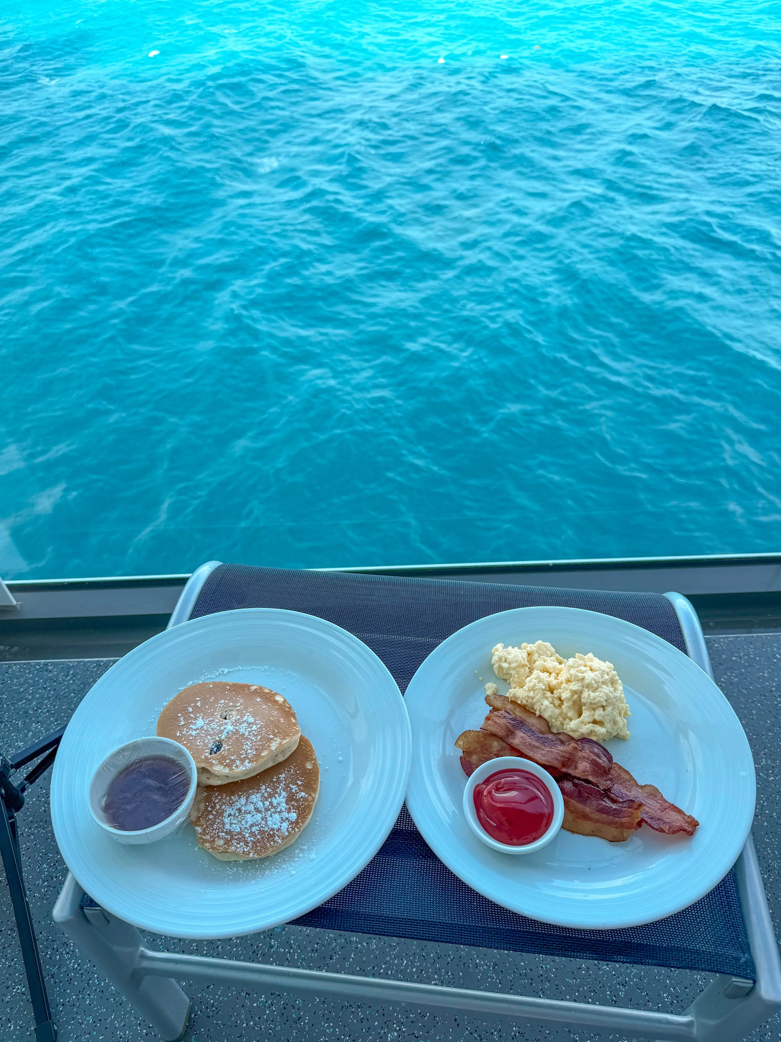 Breakfast plates with pancakes, scrambled eggs, bacon, and syrup on a table on a boat with a view of blue water outside.