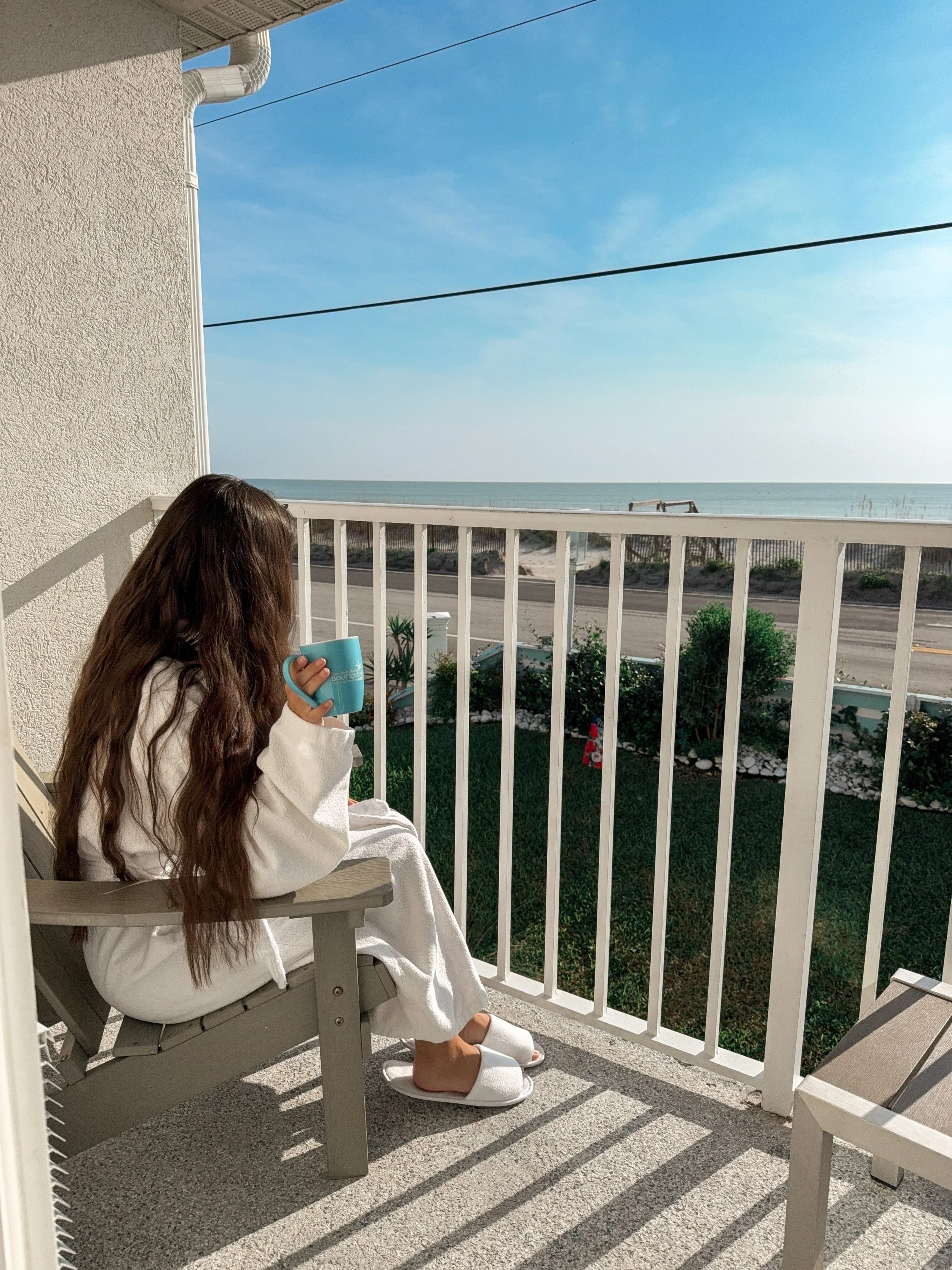 A woman with long brown hair wearing a white robe, sitting on a porch chair with a towel over her lap, holding a blue mug, with a scenic view of the ocean and a clear blue sky in the background.