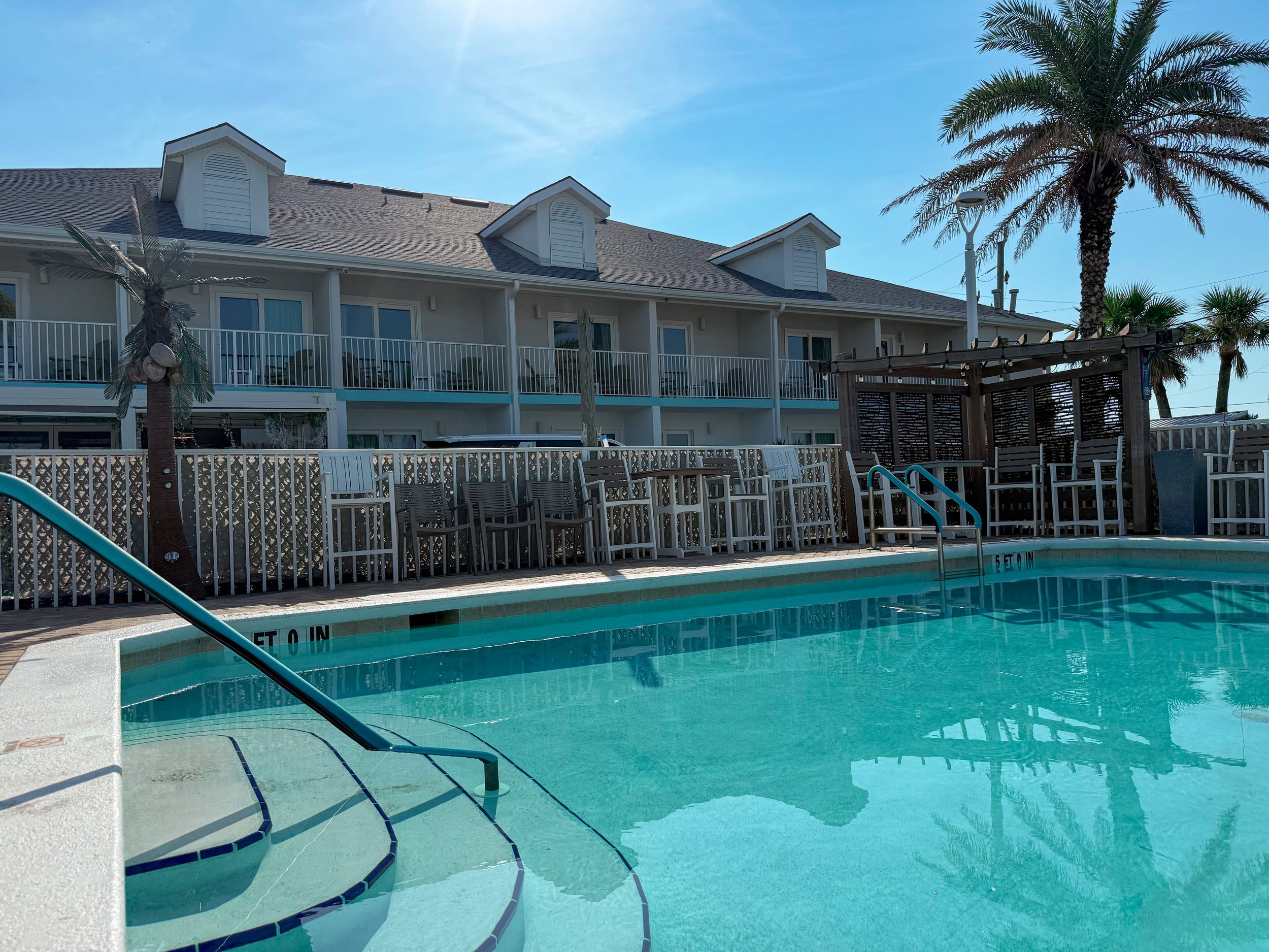 Swimming pool with steps leading into the water, surrounded by chairs, a wooden pergola, and tall palm trees, with an apartment building in the background under a clear blue sky.