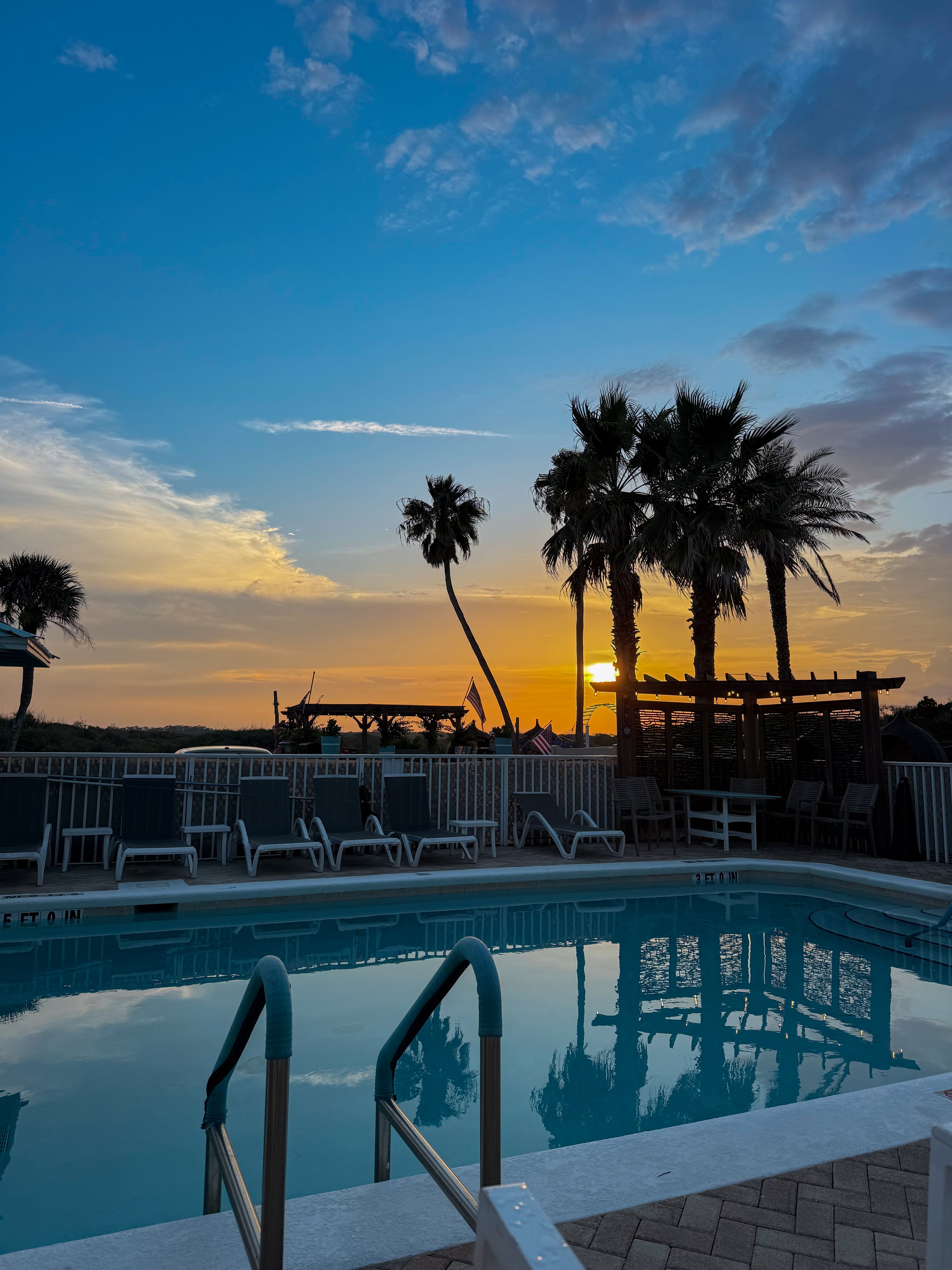 A swimming pool at sunset with palm trees in the background, lounge chairs around the pool, and a partly cloudy sky with the sun setting behind the trees.
