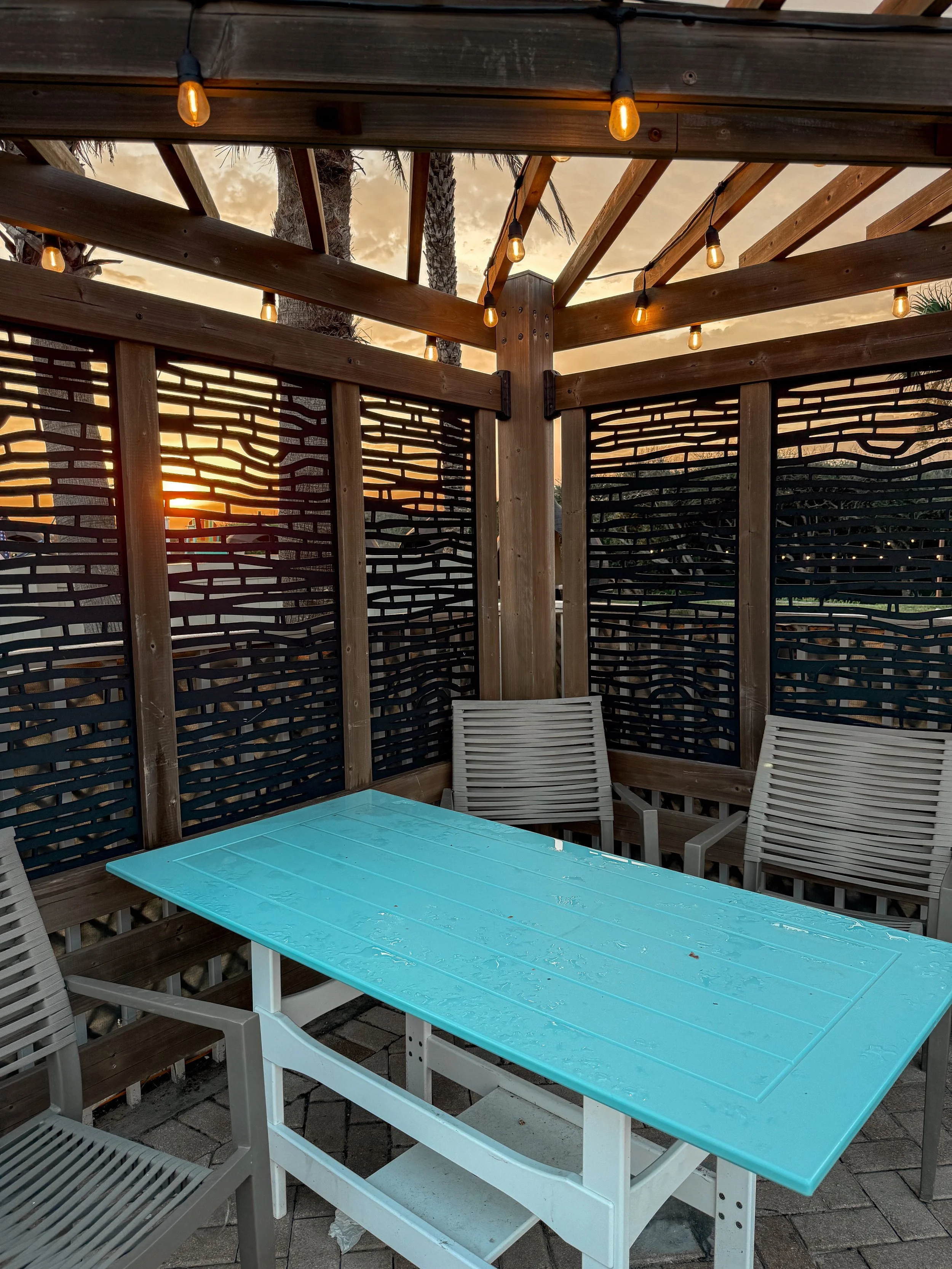 Outdoor patio area with wooden fence and string lights, a turquoise table, and plastic chairs at sunset.