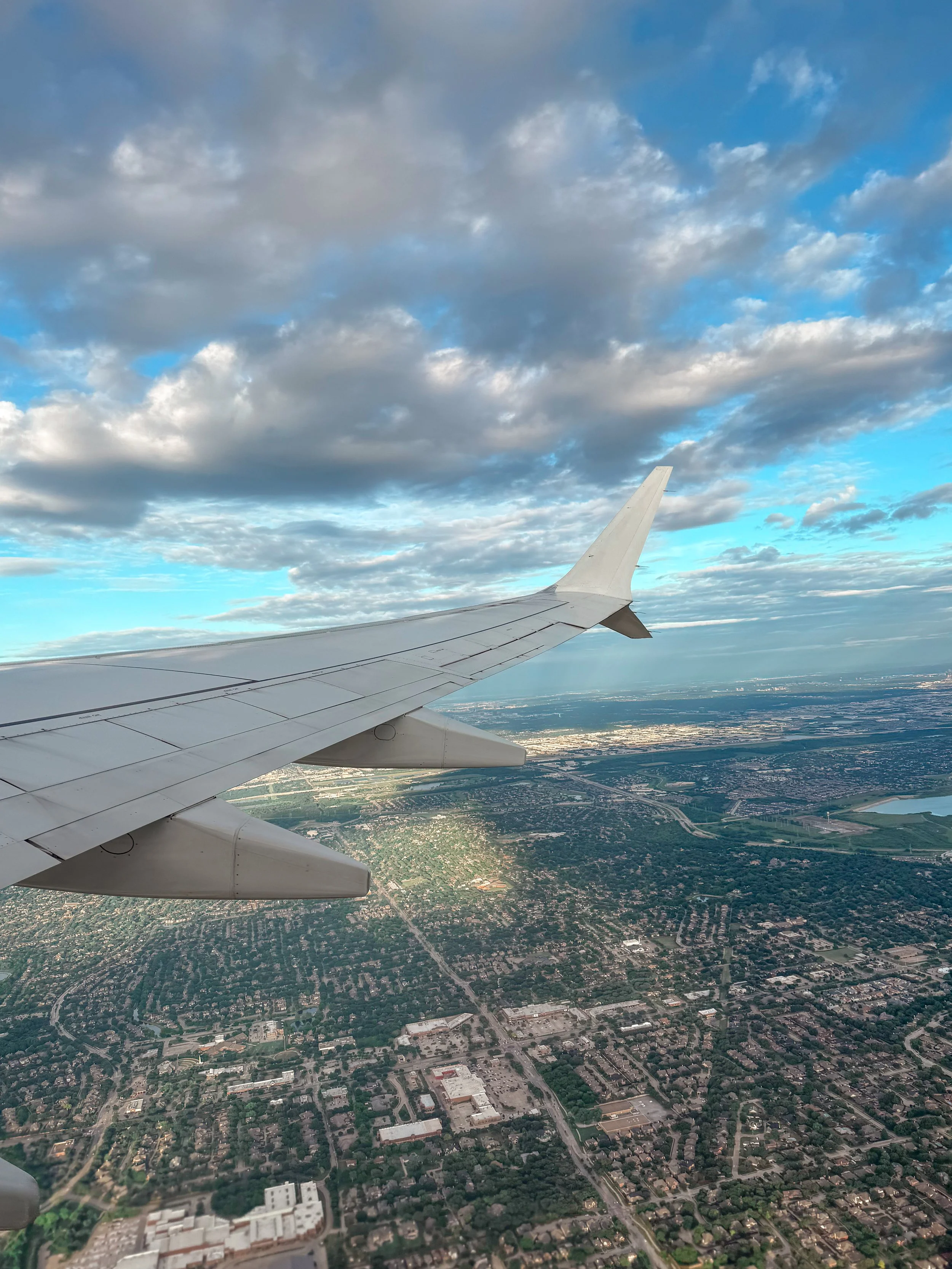 View from an airplane window showing the airplane wing, clouds in the sky, and a landscape below including roads, buildings, and green areas.