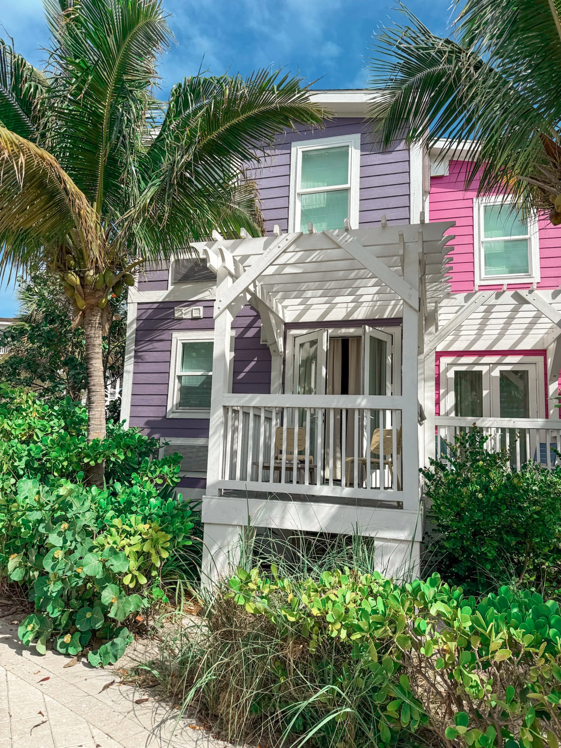 Colorful two-story house with purple and pink exterior, white trim, surrounded by lush green foliage and palm trees, with a white wooden pergola and chairs outside.