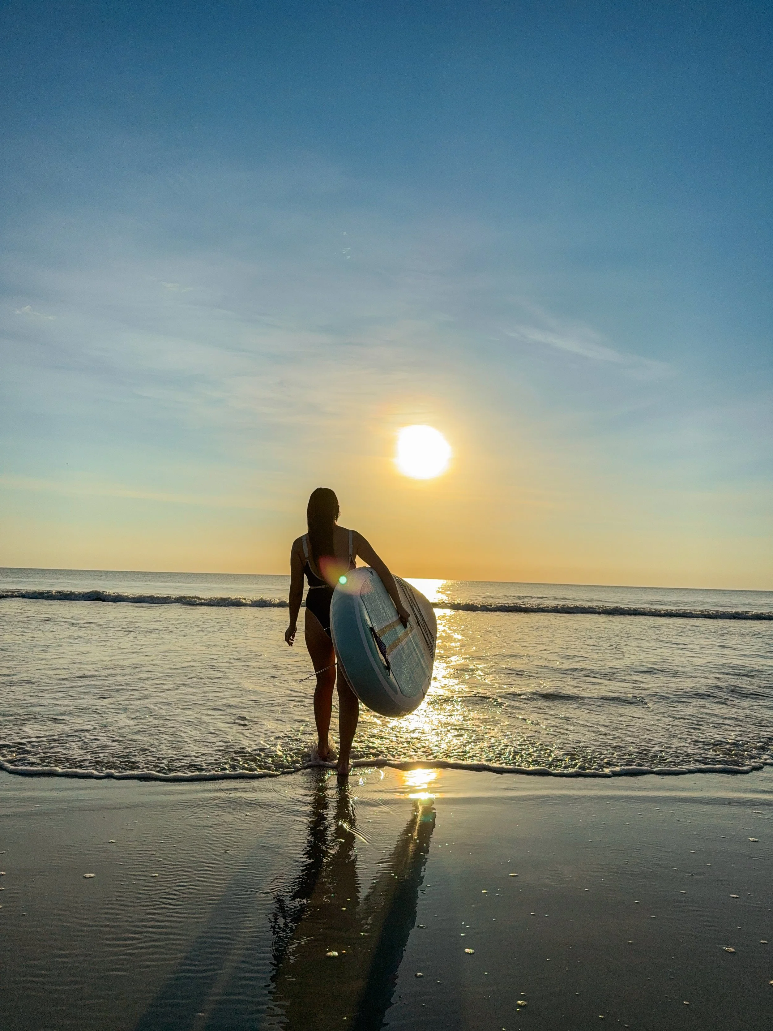 A woman walking towards the ocean with a surfboard at sunset, with the sun low in the sky and reflecting on the water.