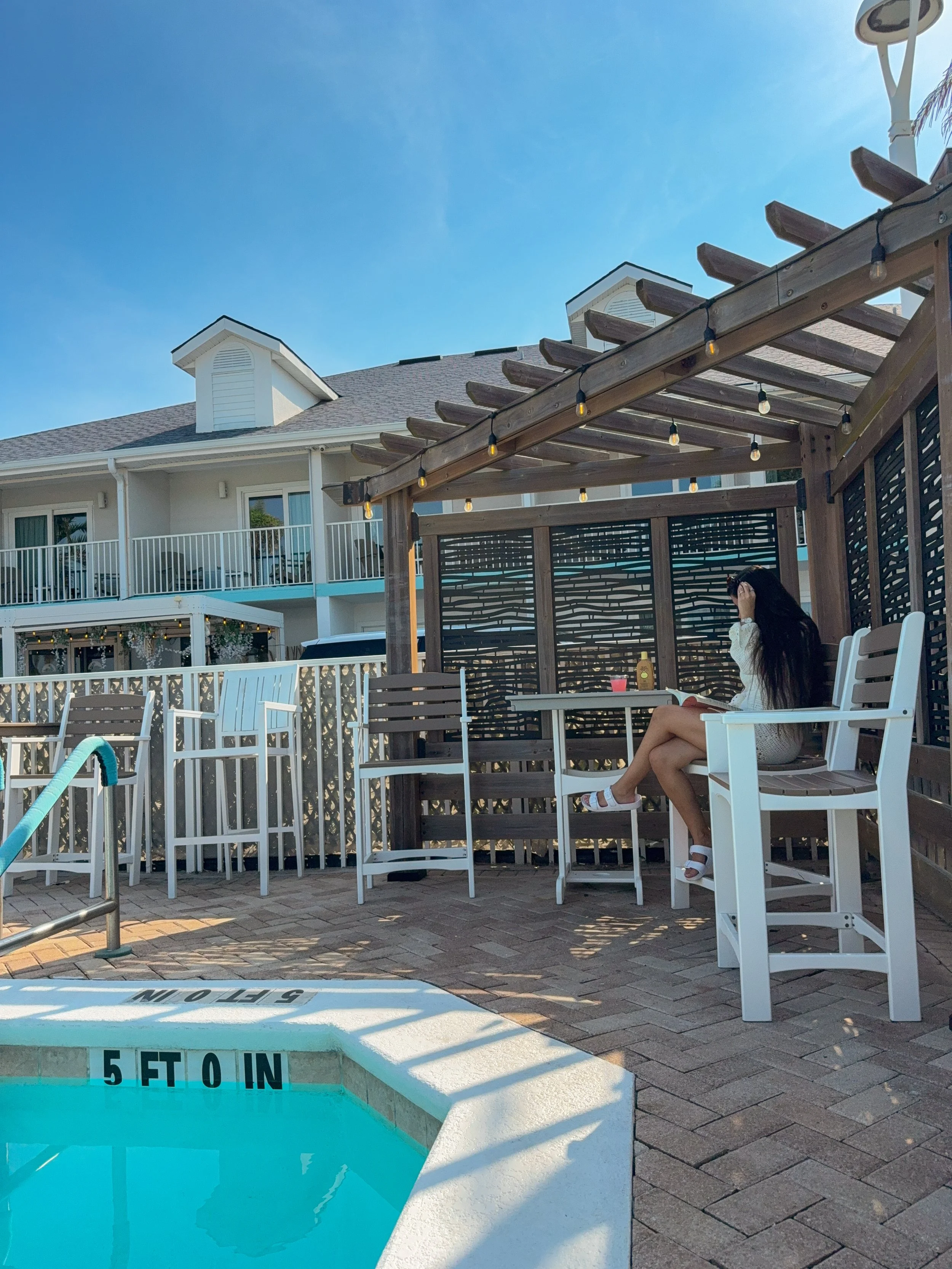Empty swimming pool near a wooden pergola, with a woman sitting on a white chair underneath, holding a phone. The pool has a depth marker of 5 feet, and the background includes a white residential building with balconies and a blue sky.