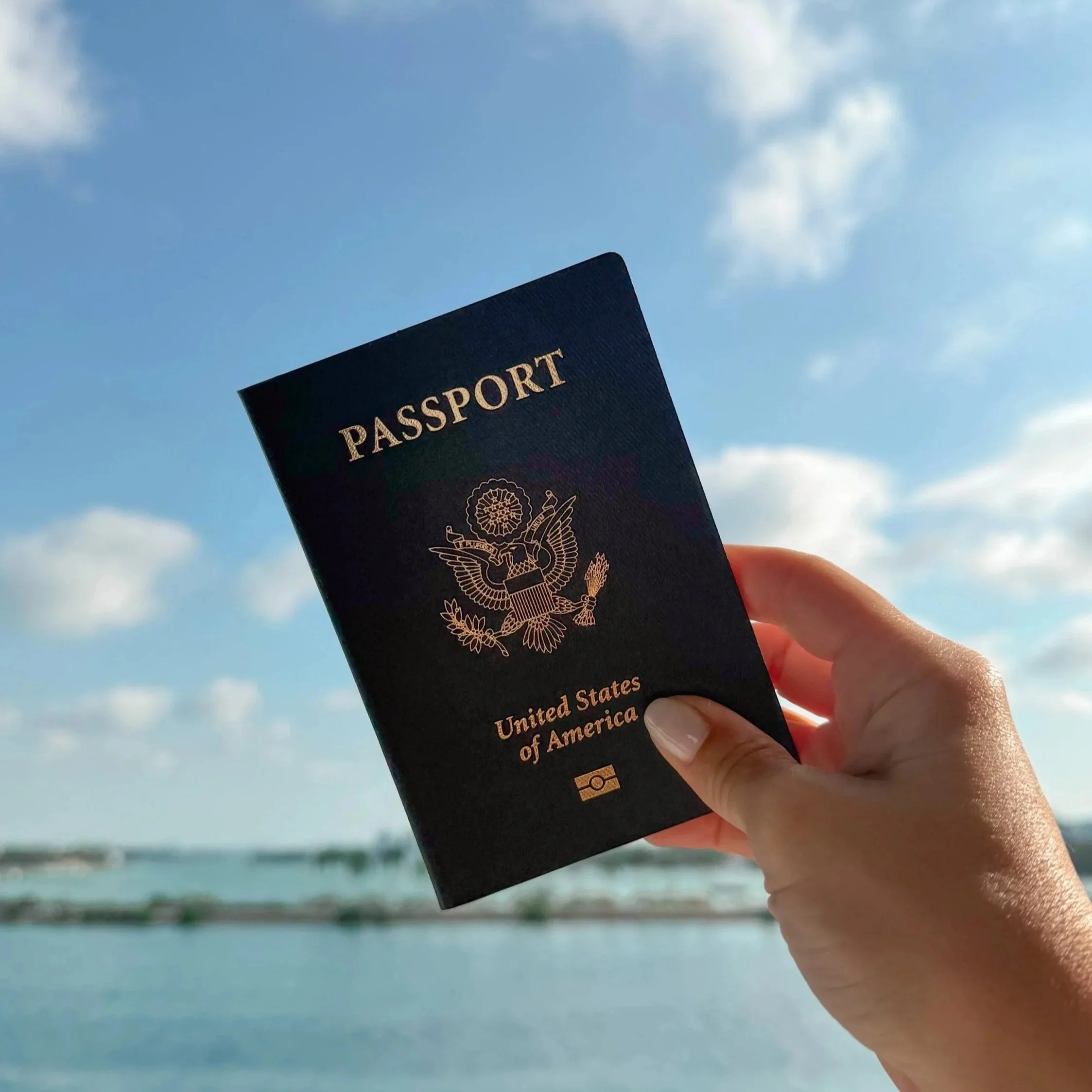 Person holding a U.S. passport against a background of blue sky with some clouds near a beach or ocean.