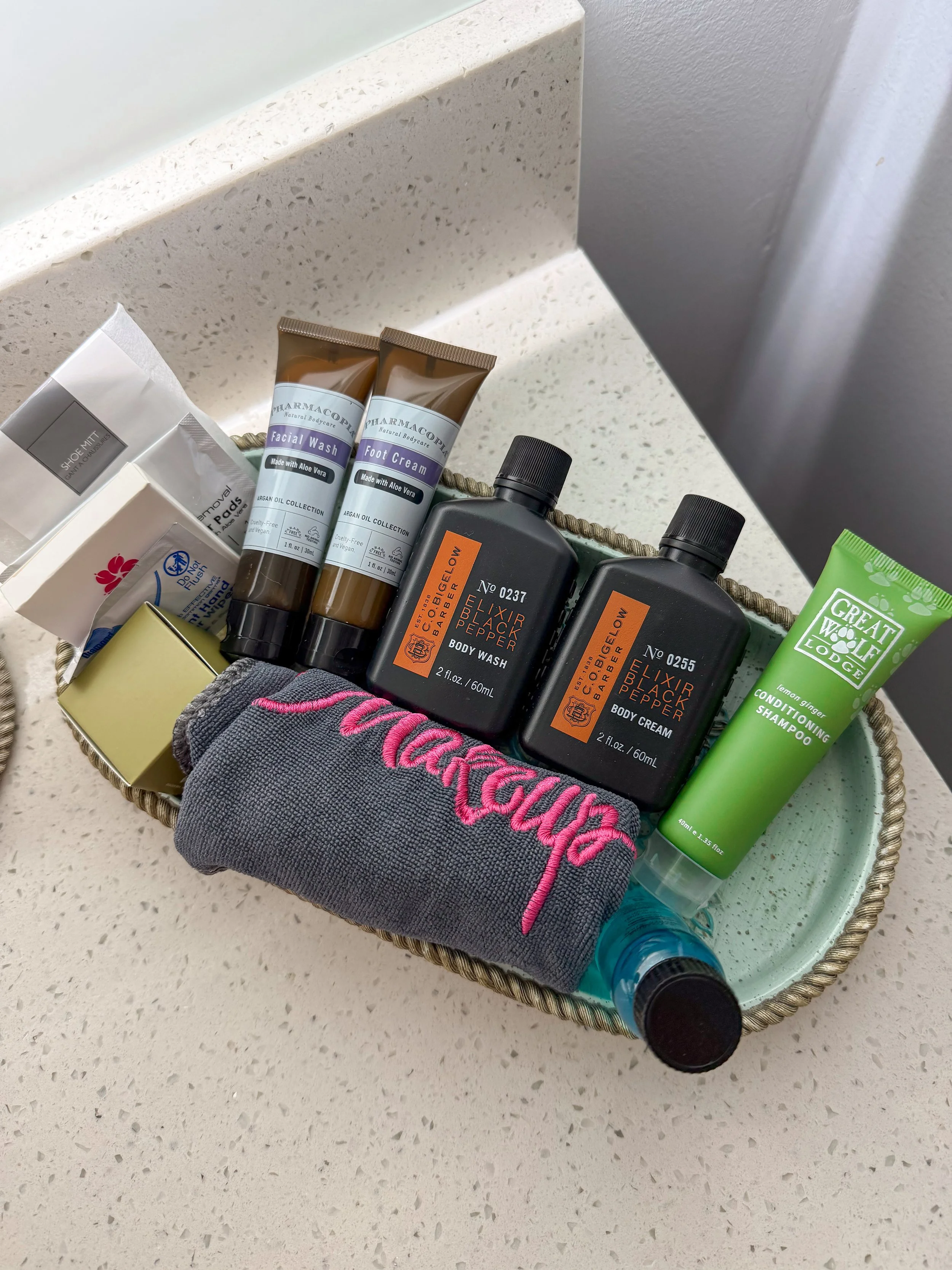 A bathroom countertop with a woven tray containing toiletries and personal care items, including two tubes labeled 'Facial Wash' and 'Foot Cream,' two bottles labeled 'Bigelow Elixir Black Pepper Body Wash' and 'Body Cream,' a green tube of 'Great Wolf Lodge Shampoo,' a pair of gray and pink socks, and a small wrapped package.